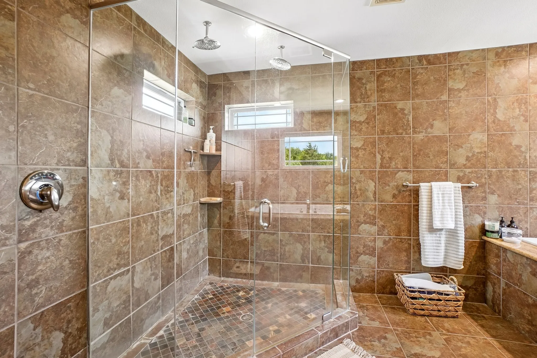 Glass-enclosed shower with multiple showerheads, natural-toned wall and floor tiling, and integrated shower niche