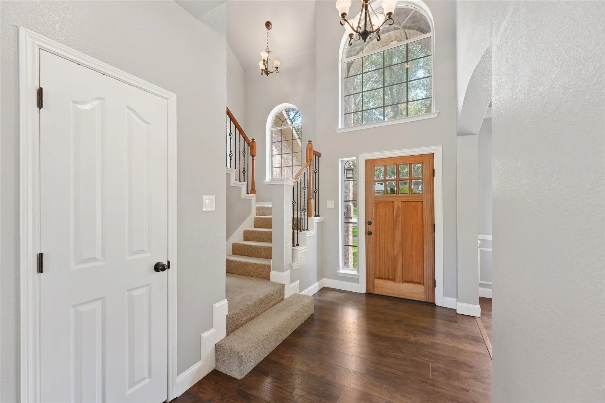 Wood-finish flooring, staircase with carpeted treads and wrought iron balusters, natural wood entry door with glass panels, arched transom window, and a chandelier lighting fixture