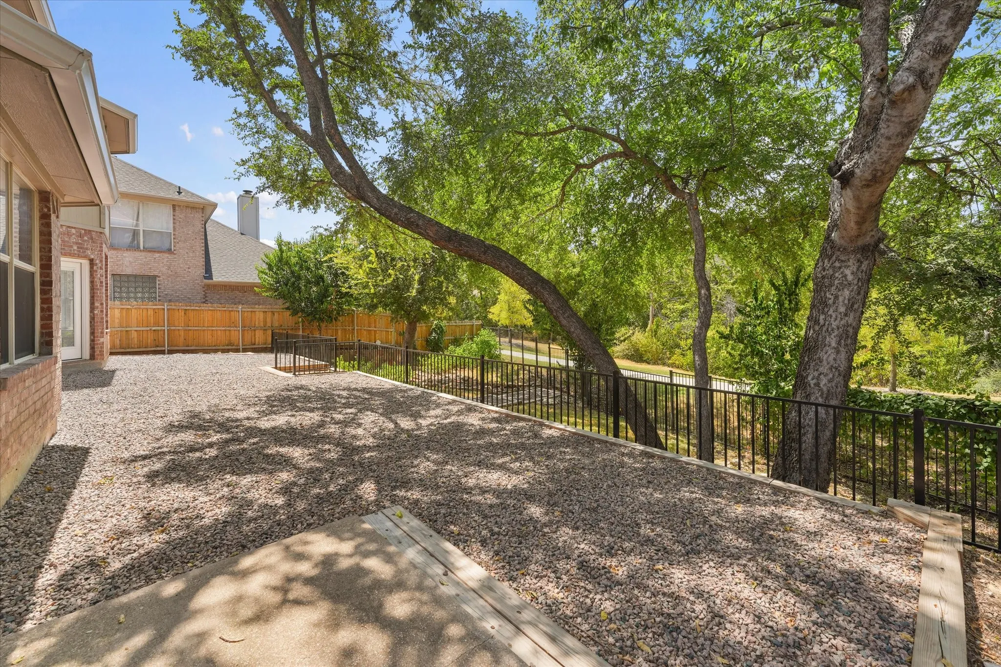 Expansive outdoor area featuring a concrete patio, gravel landscaping, mature trees, and a black metal fence