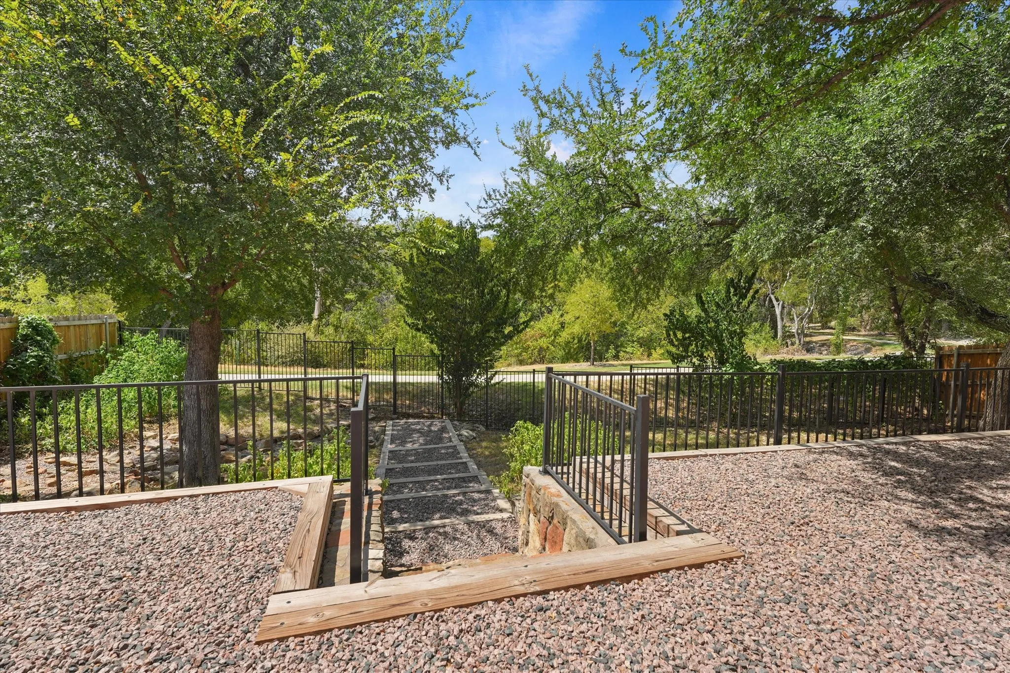 Gravel-covered outdoor area with stone steps, black metal fencing, and mature trees