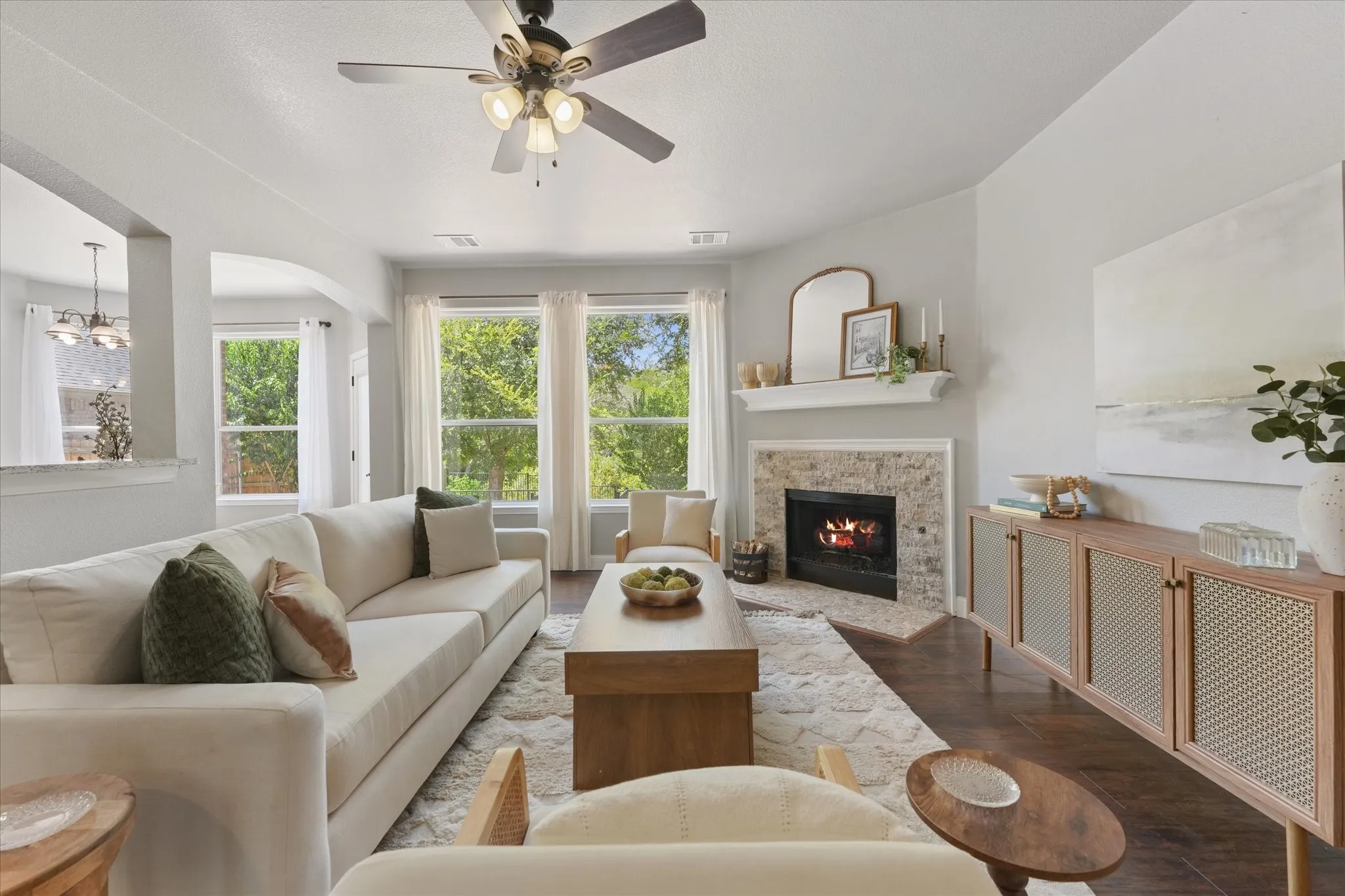 Vaulted ceiling with a ceiling fan, stone-clad corner fireplace with a mantel, dark wood-finish flooring, and multiple large windows