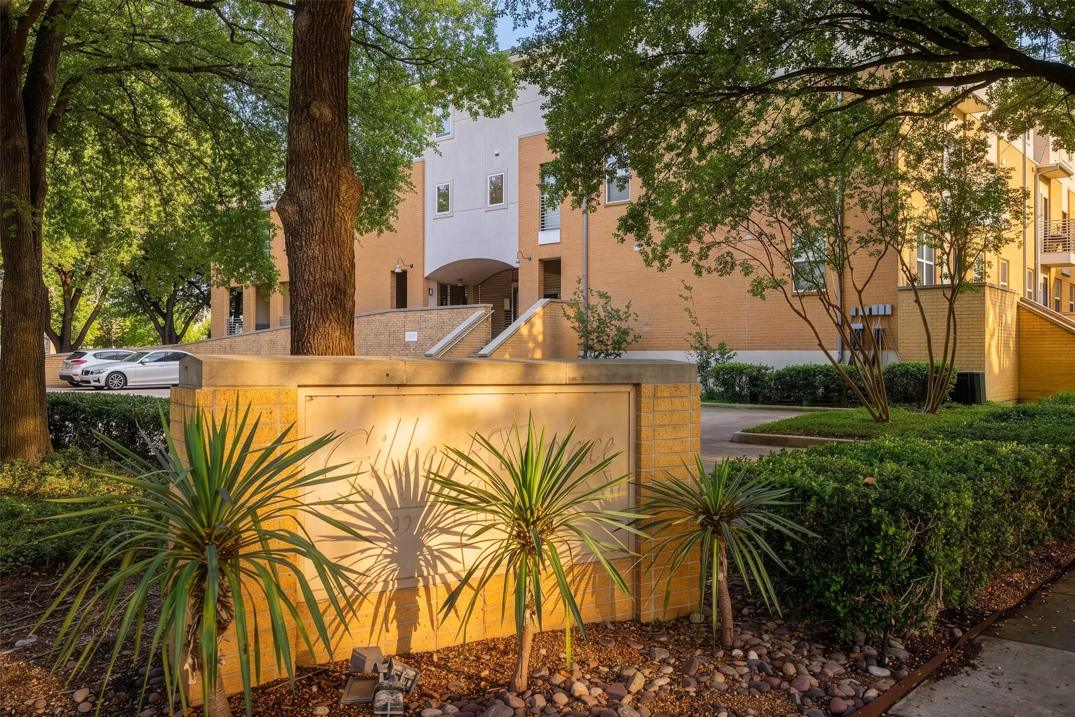 Multi-story building exterior featuring brick and stucco finishes, mature trees, manicured shrubs, and a landscaped entrance with a stone-clad retaining wall
