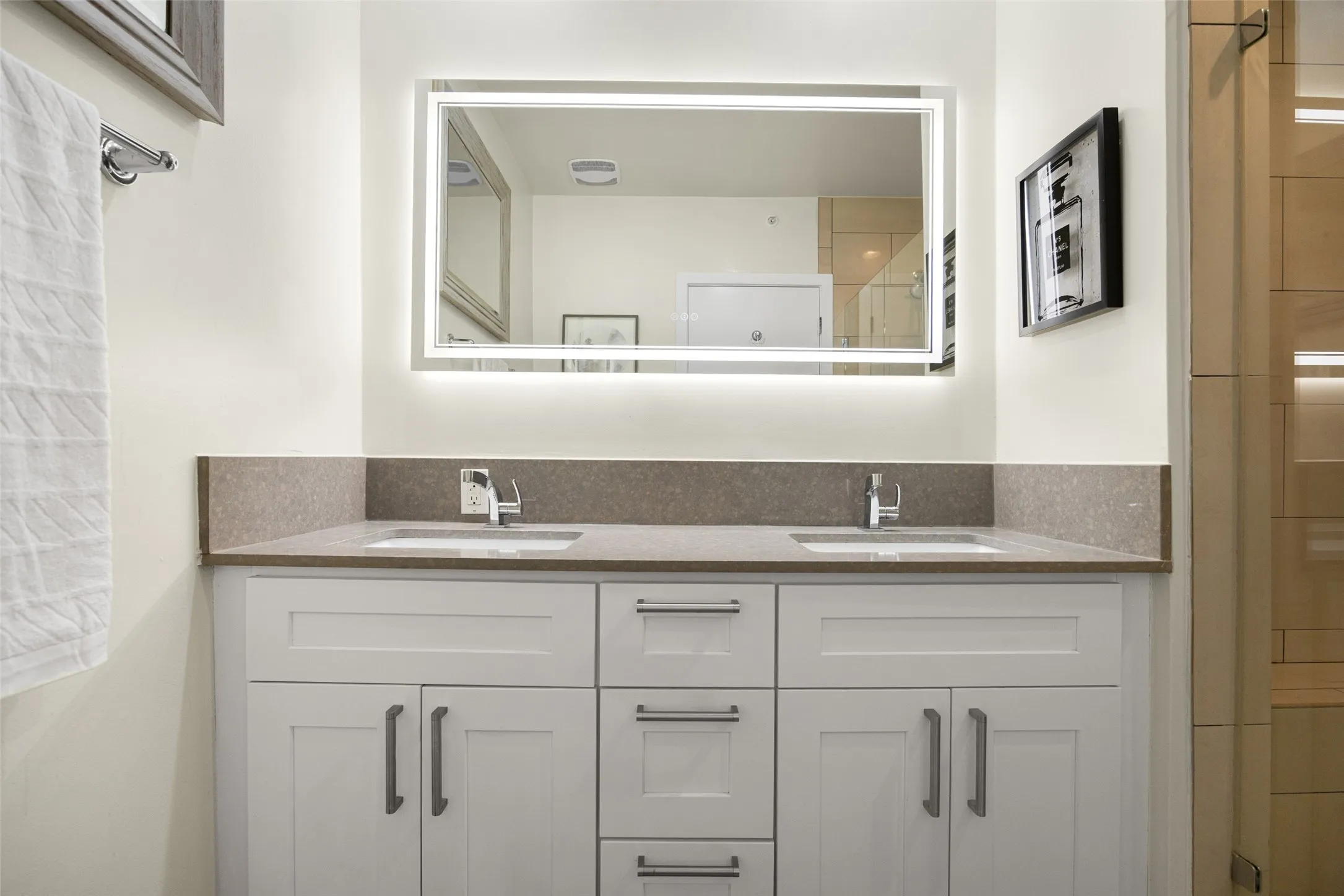 Dual vanity with integrated sinks, a stone countertop, white shaker-style cabinetry, and a backlit​​‌​​​​‌​​‌‌​‌‌​​​‌‌​‌​‌​‌​​​‌​​ mirror