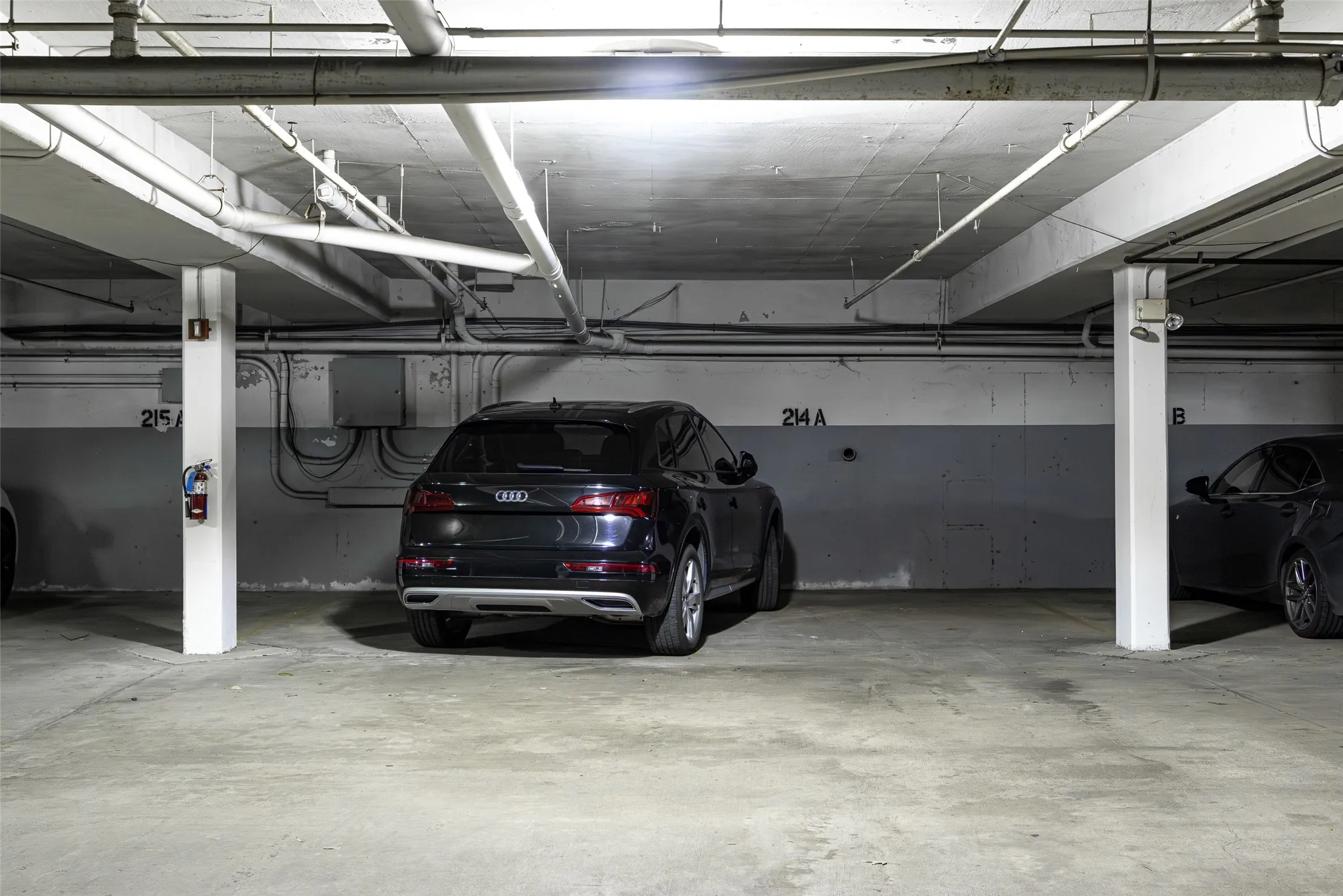 Designated parking space within a concrete garage structure, featuring exposed overhead piping, concrete columns, and wall-mounted electrical conduits