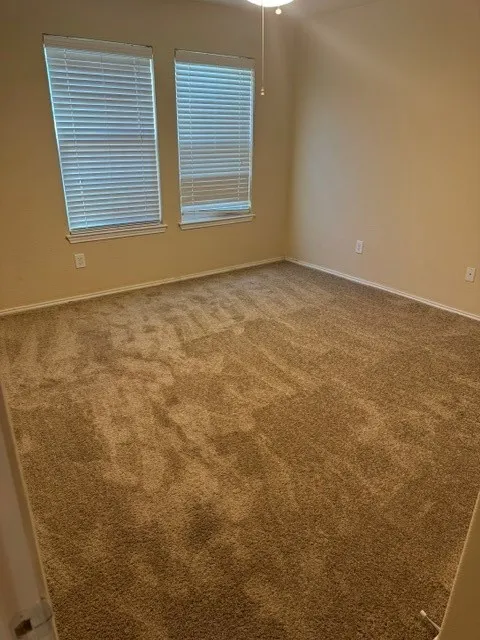 Neutral-toned carpeting throughout, featuring two windows with horizontal blinds, and a ceiling fan with integrated lighting