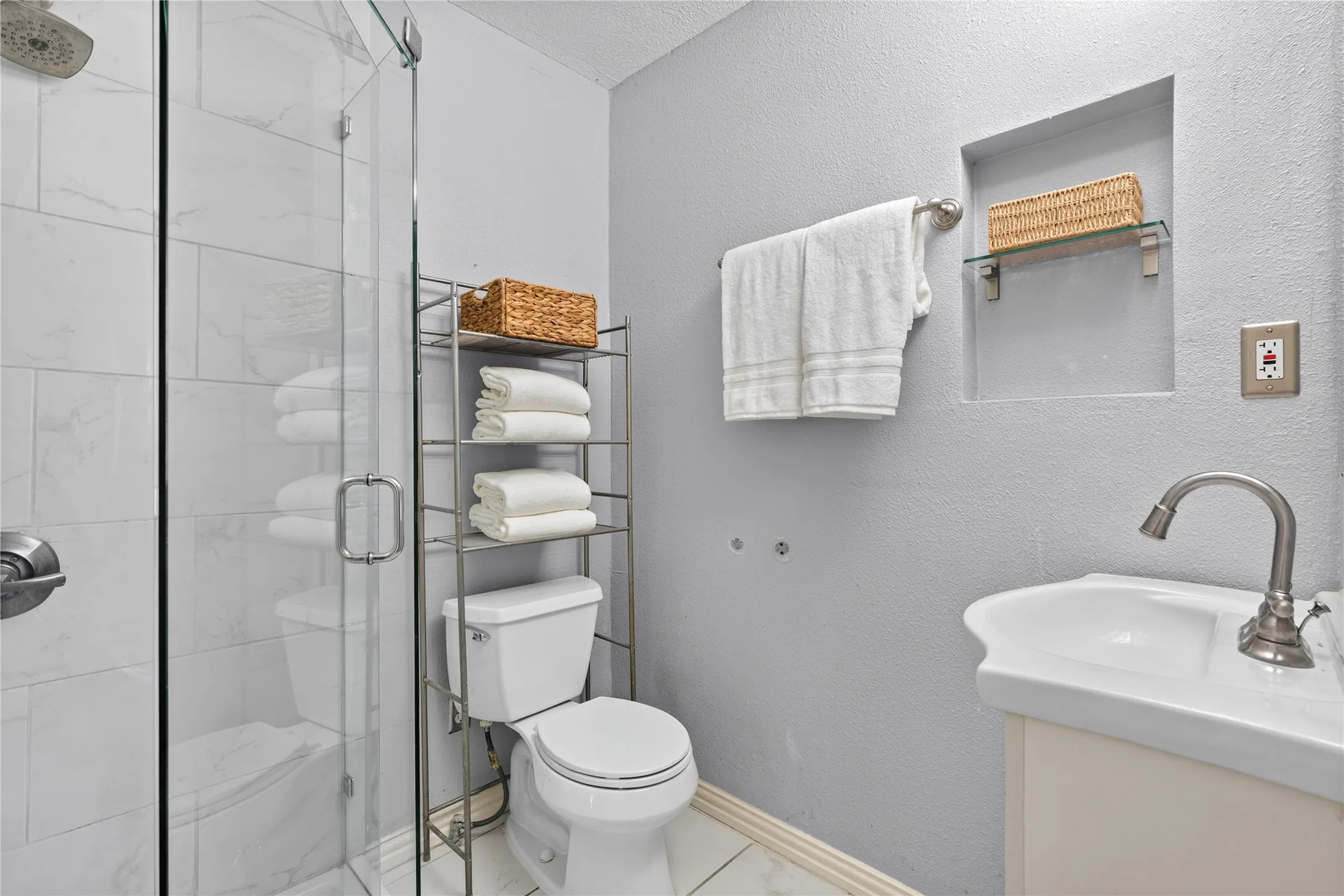 Glass-enclosed shower with subway tile surround, white pedestal sink with brushed nickel faucet, built-in wall niche with glass shelf, and a white toilet