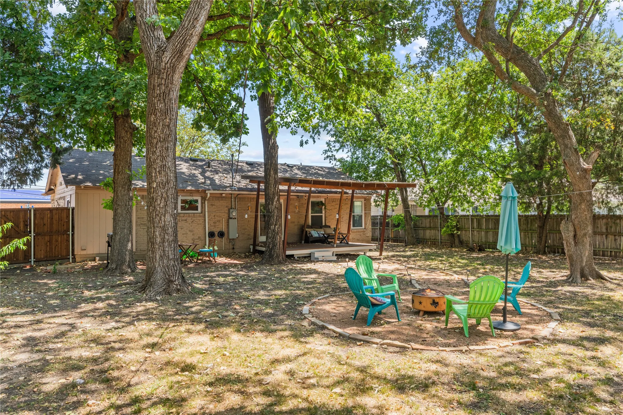 Expansive backyard featuring mature trees, a wooden privacy fence, a covered patio with a wood-finish ceiling, and a built-in outdoor fire pit area