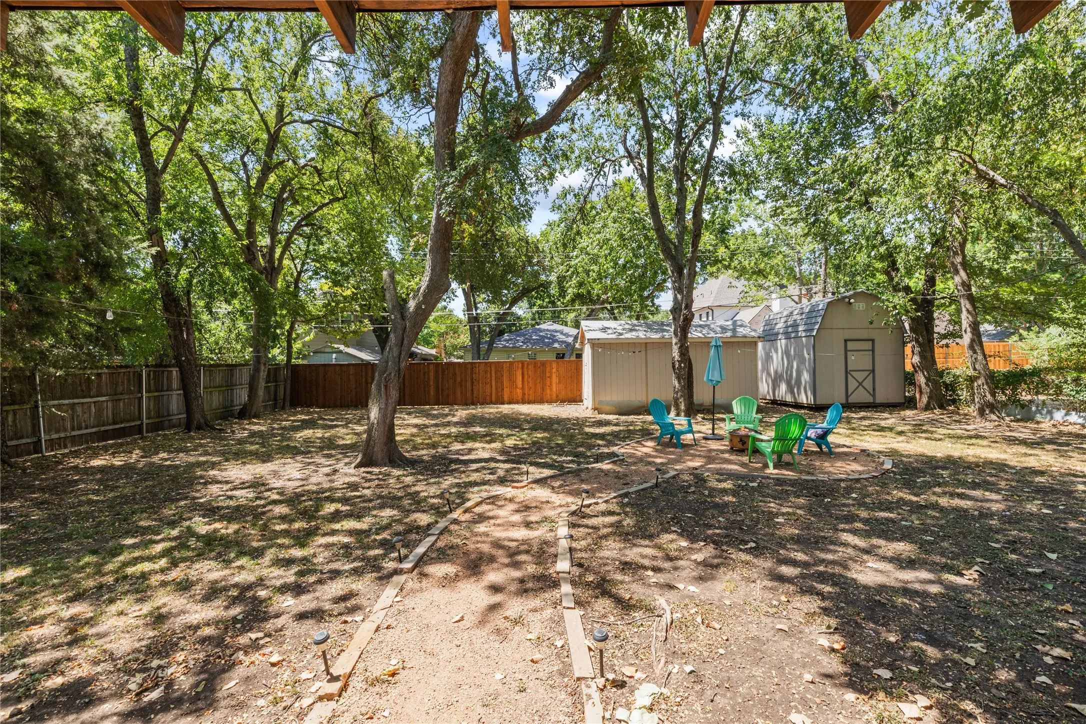 Expansive backyard with a full privacy fence, mature trees, a dedicated fire pit area, and two storage sheds