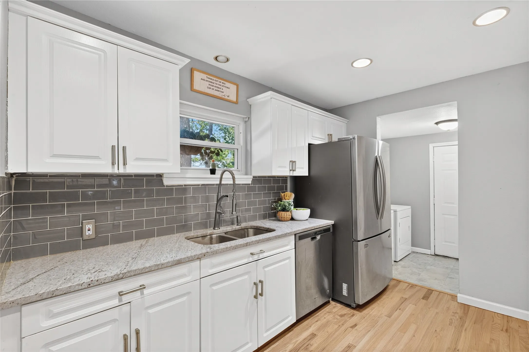 White cabinetry with brushed nickel hardware, gray subway tile backsplash, light-toned countertop, dual basin stainless steel sink, and wood-finish flooring