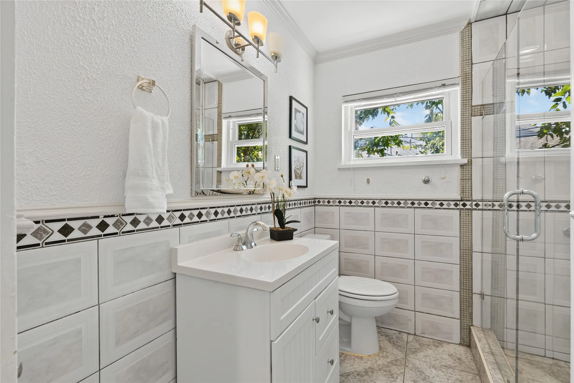 White vanity with countertop sink, framed mirror with sconce lighting, tiled walls with decorative border, and a glass-enclosed shower