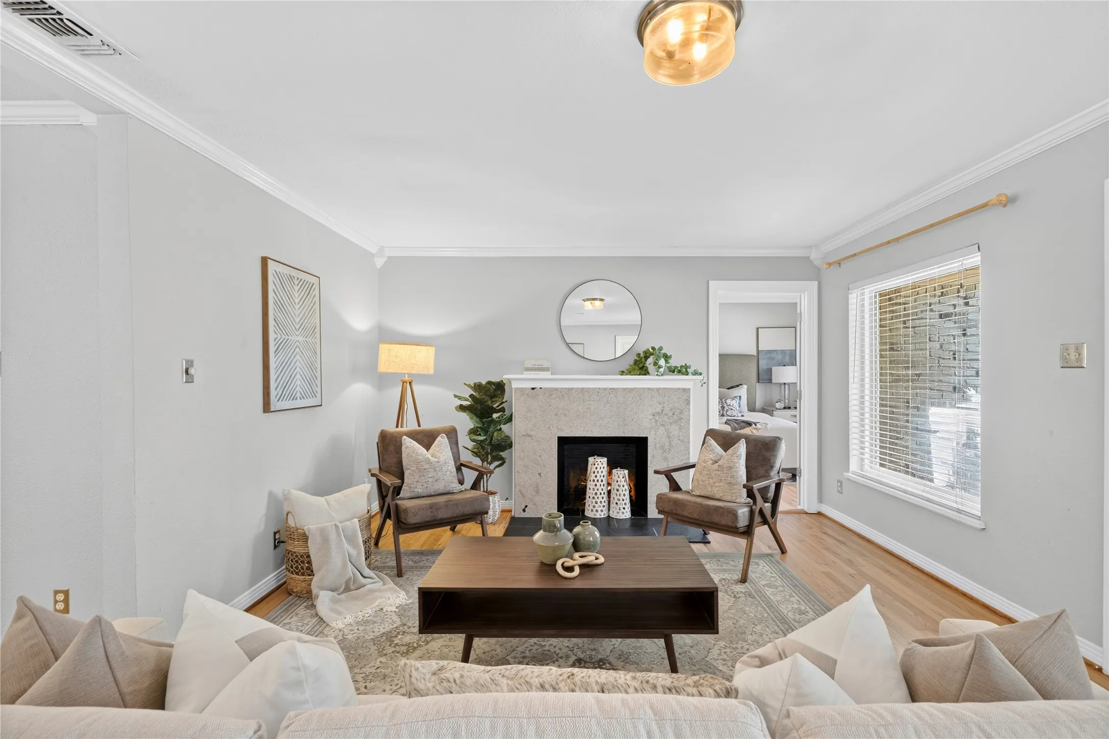 Living area featuring hardwood flooring, a stone-look fireplace, crown molding, and a prominent window with blinds