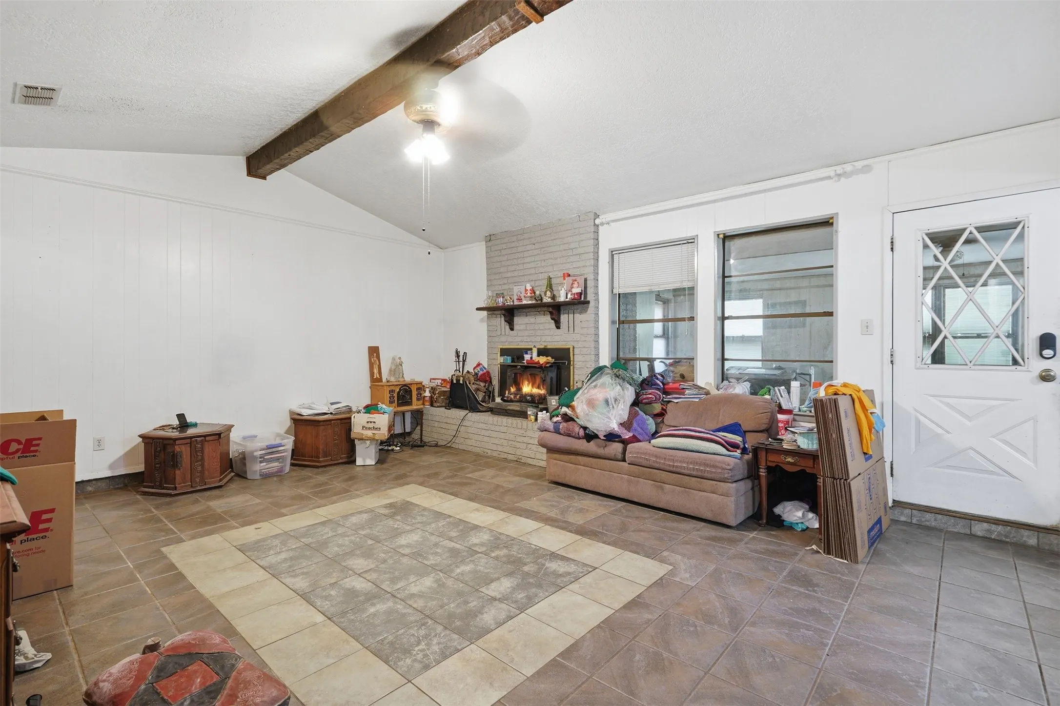 Vaulted ceiling with an exposed wood beam, ceiling fan, brick fireplace with mantel, white paneling, and tile flooring with a decorative inlay