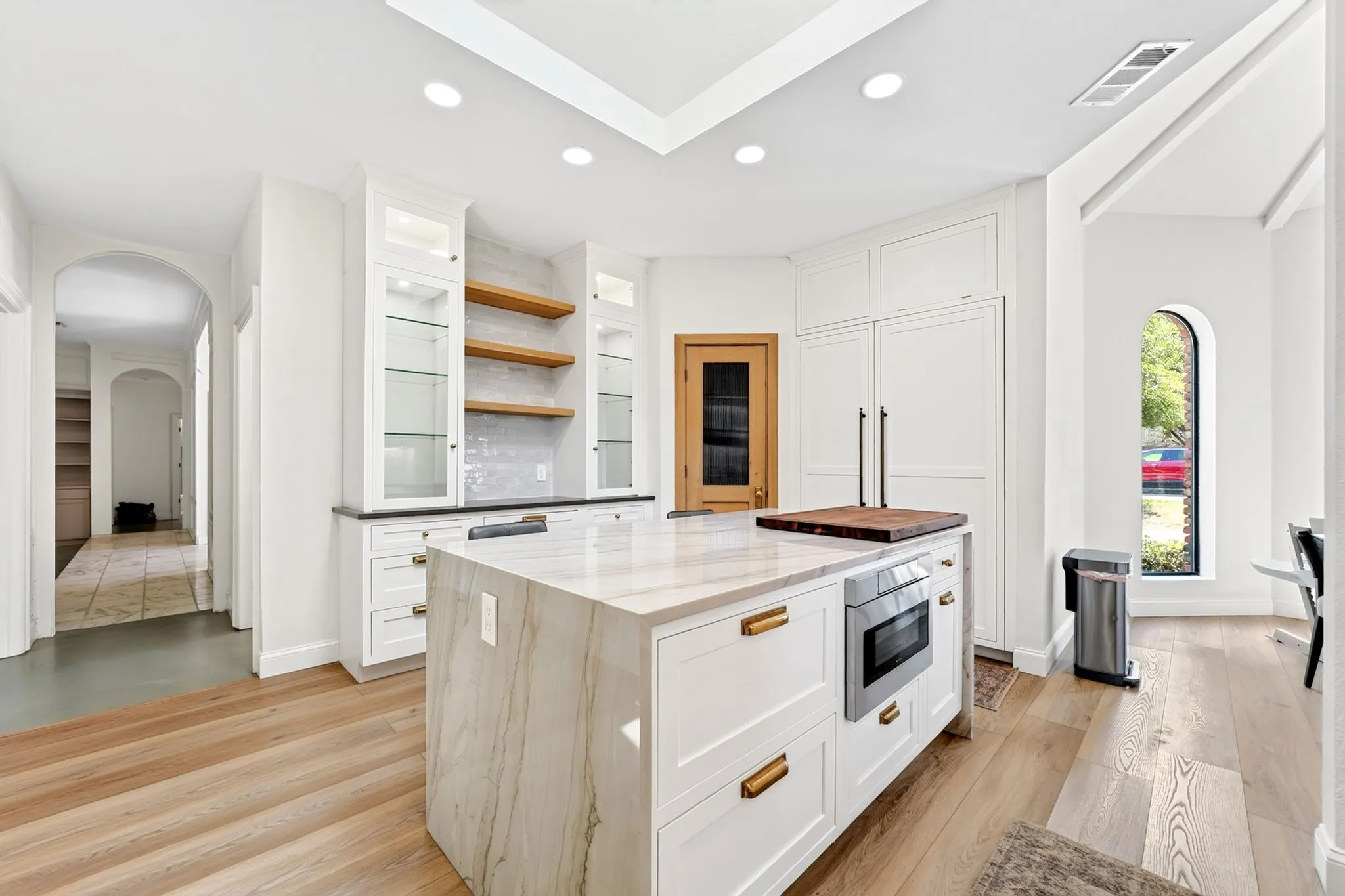 Spacious kitchen featuring a large island with stone countertop, wood-finish flooring, built-in microwave, white cabinetry with glass display doors, and an arched window