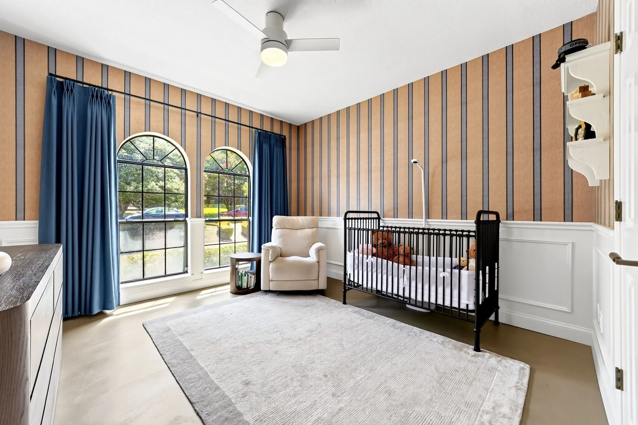 Dual arched windows with dark muntins, decorative wainscoting, striped wall covering, and a ceiling fan with integrated lighting