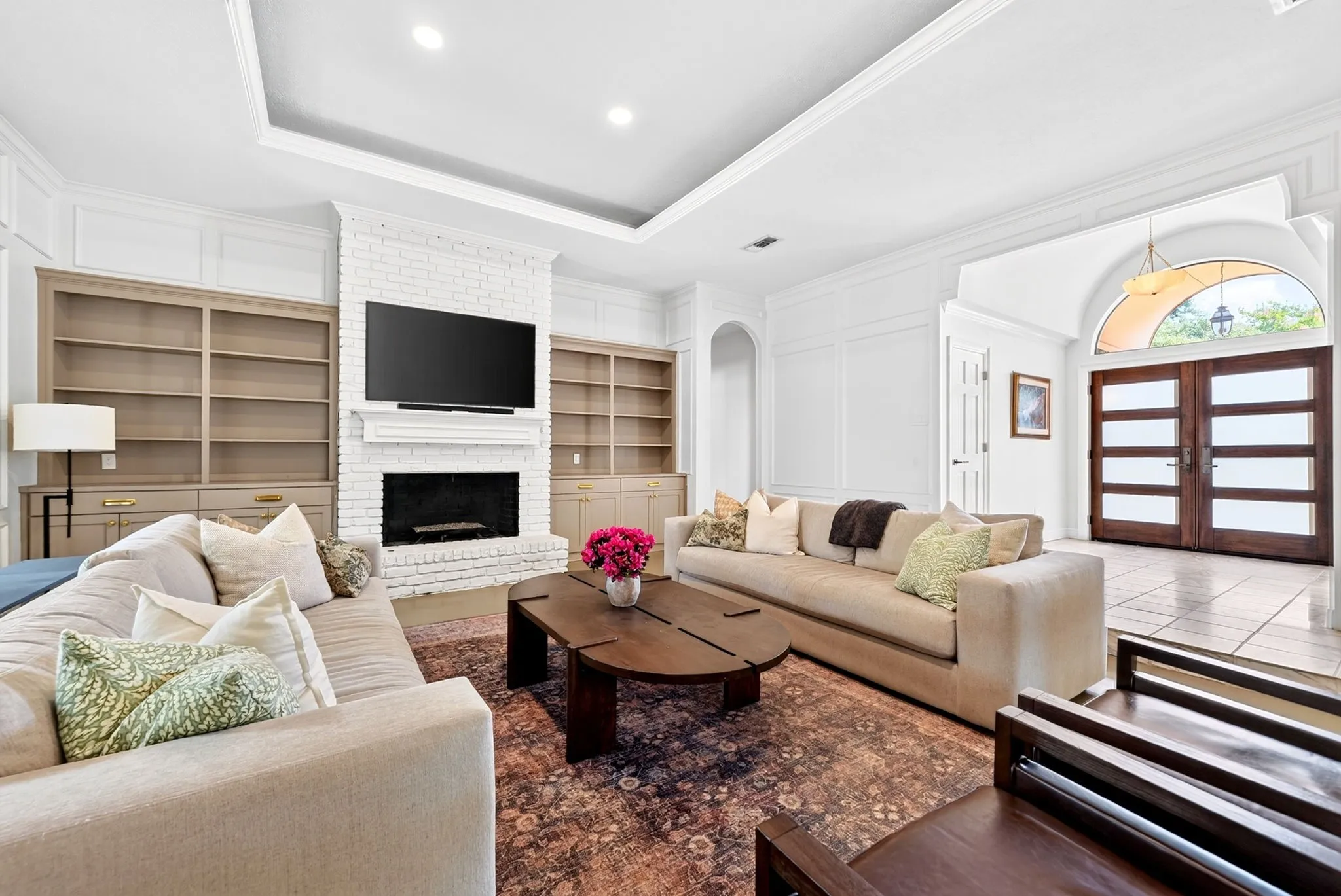 White brick fireplace with mantel, tray ceiling with recessed lighting, built-in shelving, wood double entry doors with frosted glass, and tile flooring