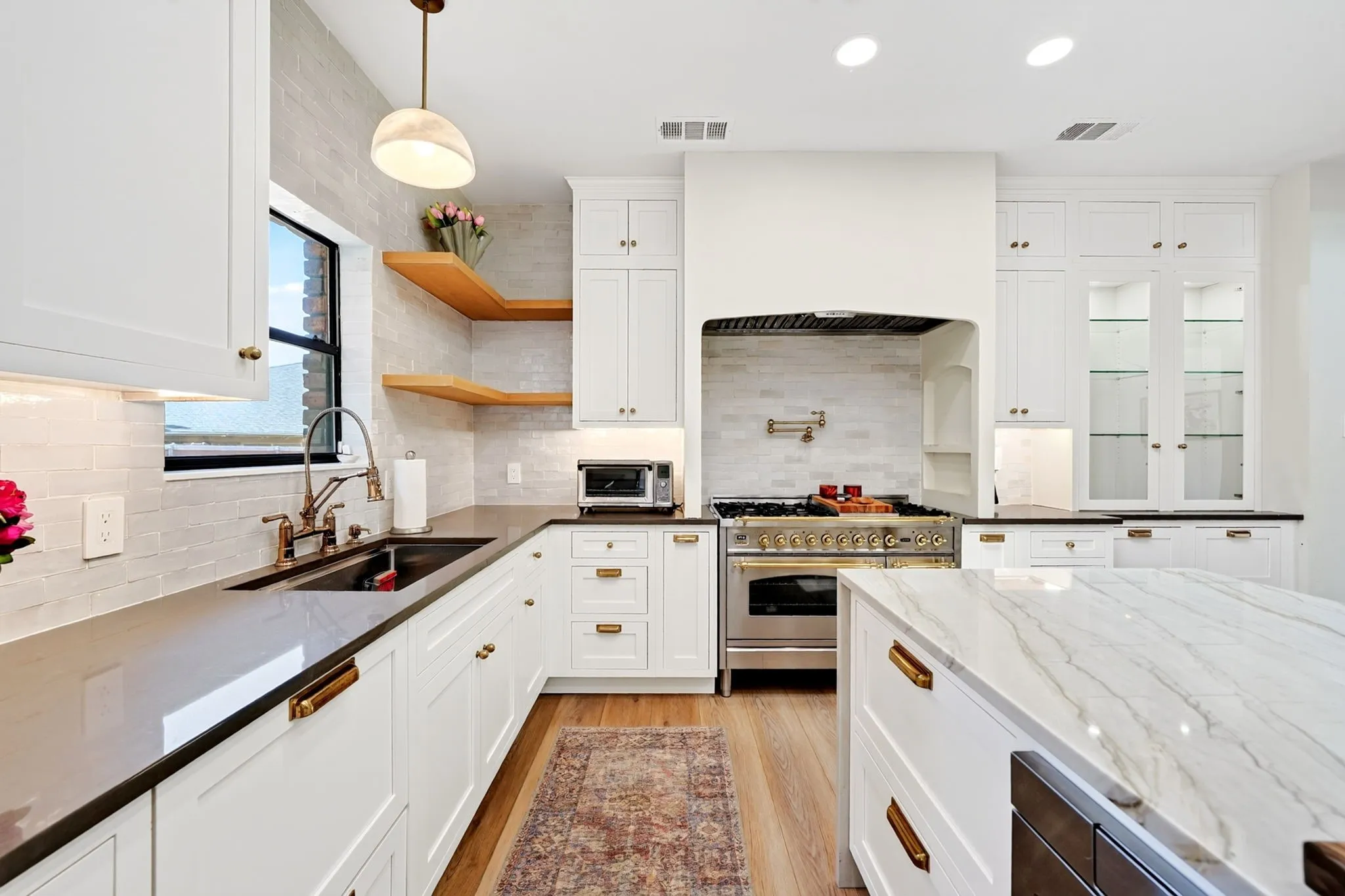 White shaker cabinetry with gold-tone hardware, wood-finish flooring, a kitchen island with a stone countertop, an undermount double-basin sink, and a stainless steel gas range