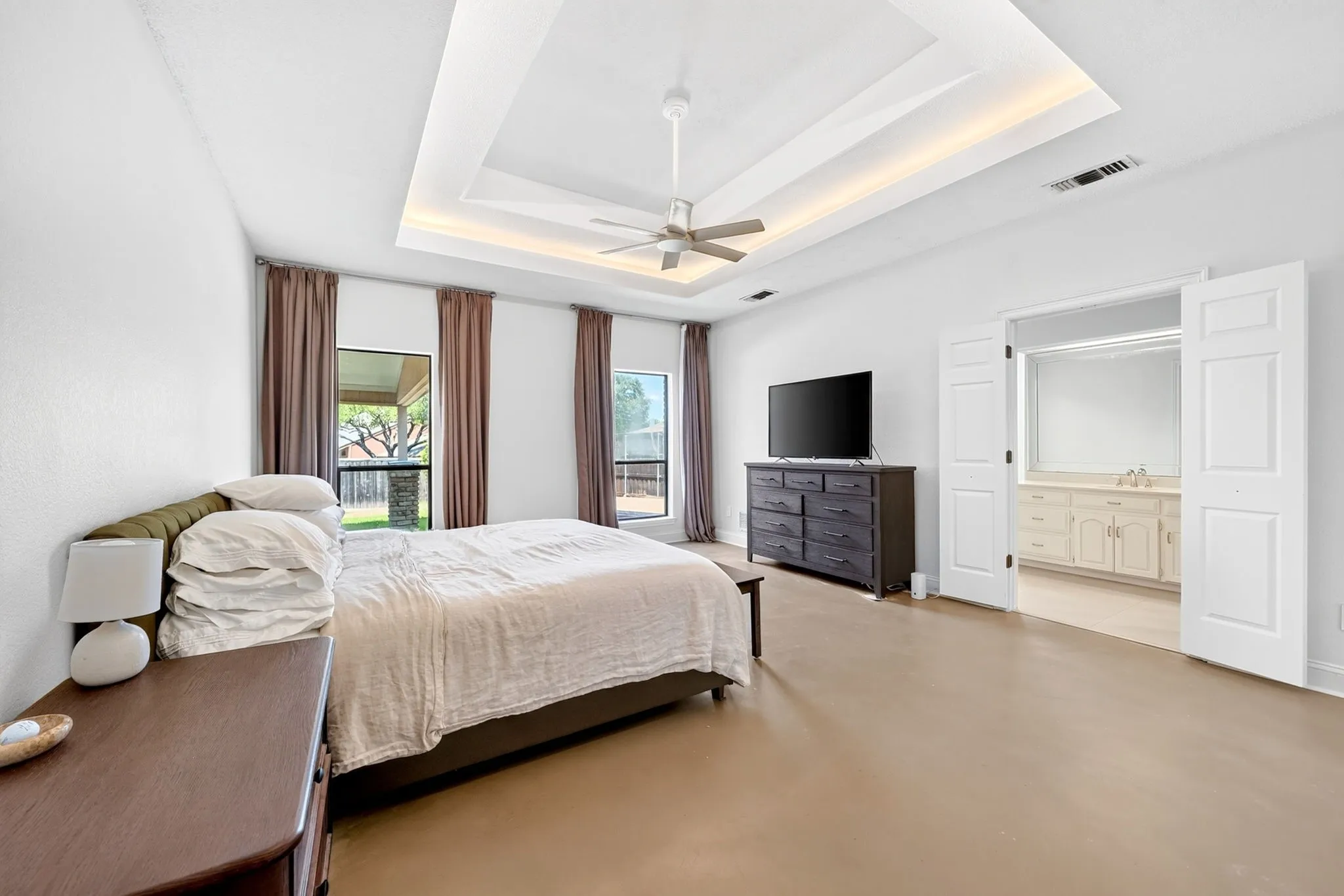 Tray ceiling with cove lighting and ceiling fan, two windows with exterior views, wide-plank flooring, and double doors opening to an ensuite bathroom