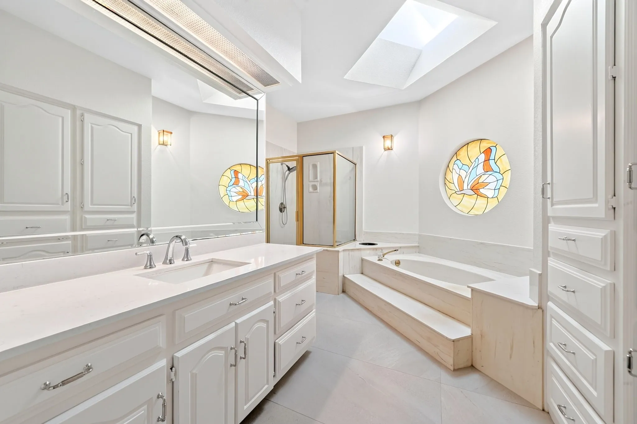Recessed lighting with a large skylight, dual vanity with a single basin sink, built-in cabinetry, and a soaking tub with stepped access