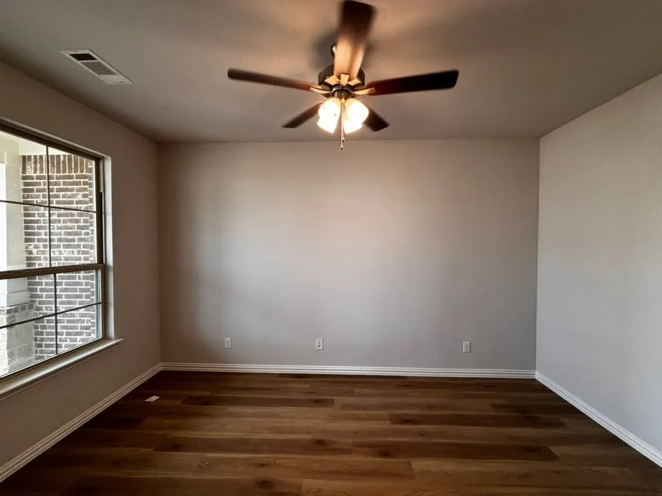 Wood-finish flooring, neutral wall paint, ceiling fan with integrated lighting, and a large window with exterior brick detailing
