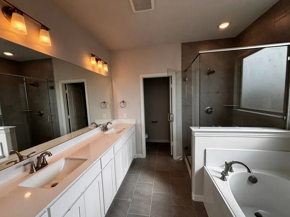 Dual vanity with integrated sinks and white cabinetry, a walk-in shower with glass enclosure and tiled surround, a separate soaking tub with deck-mounted fixtures, and tile flooring