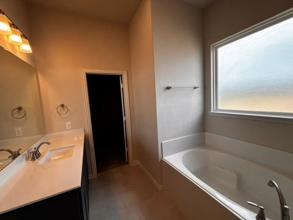 Built-in soaking tub with a frosted window, dual vanity with undermount sinks, light-toned countertop, and tile flooring