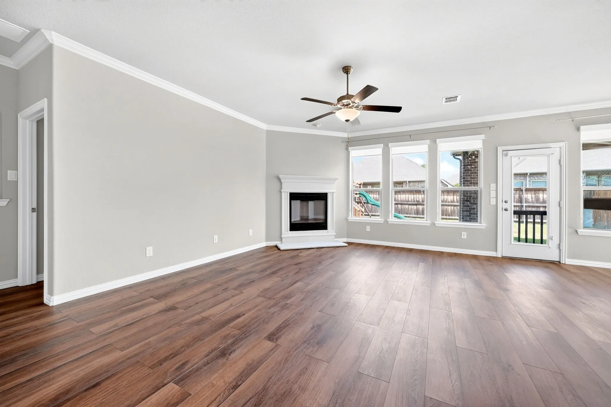 Spacious room featuring wood-finish flooring, a white mantel fireplace, multiple windows with roller shades, and a ceiling fan