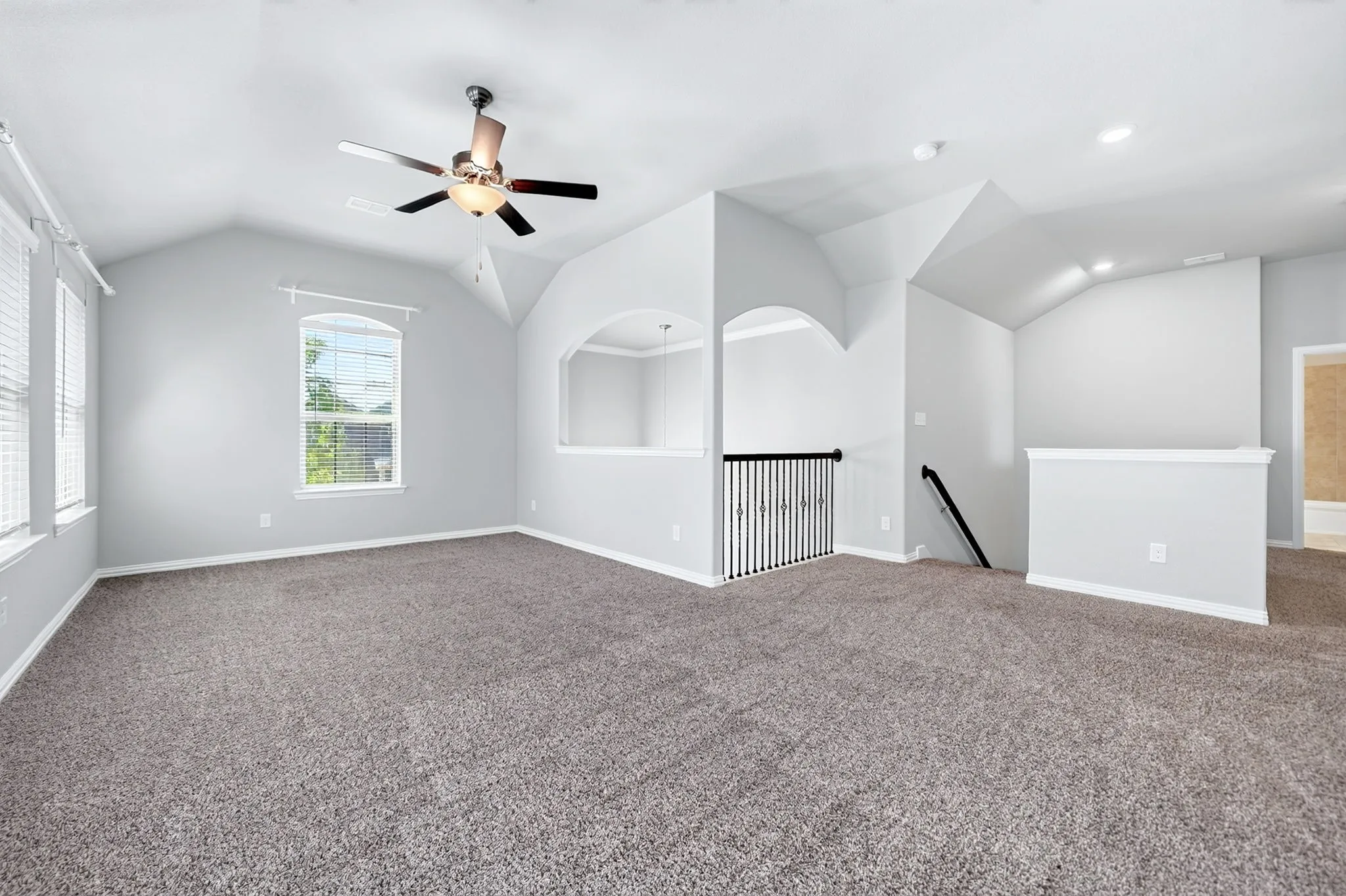 Spacious carpeted room featuring a vaulted ceiling with a ceiling fan, multiple windows with blinds, and a built-in arched alcove overlooking a lower level