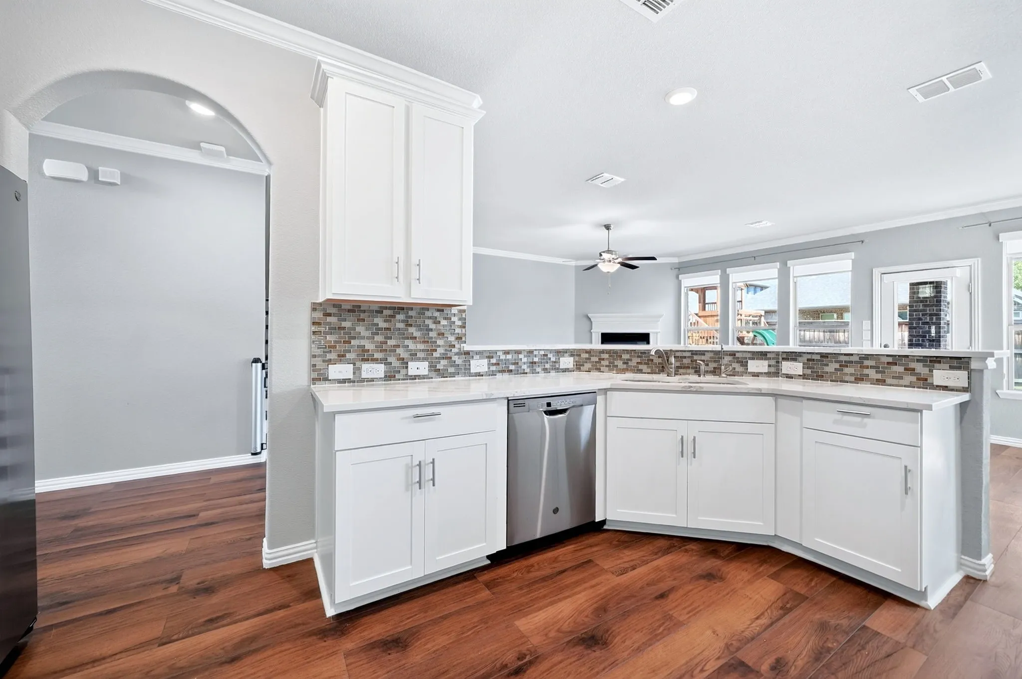 White cabinetry with brushed nickel hardware, stainless steel dishwasher, mosaic tile backsplash, solid surface countertops, and wood-finish flooring