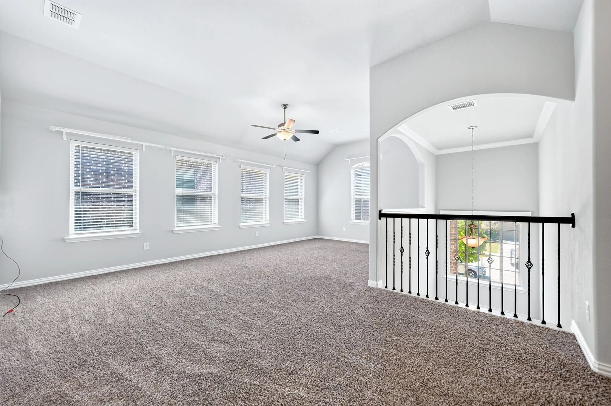 Spacious carpeted area featuring a ceiling fan, multiple windows with blinds, a wrought iron railing, and an arched opening