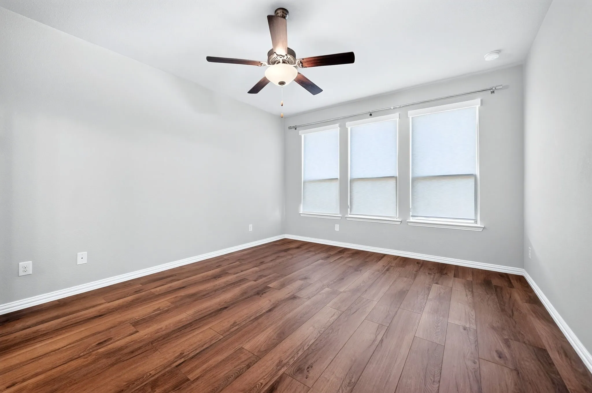 Wood-finish flooring with white trim, a ceiling fan with light fixture, and three windows with roller shades