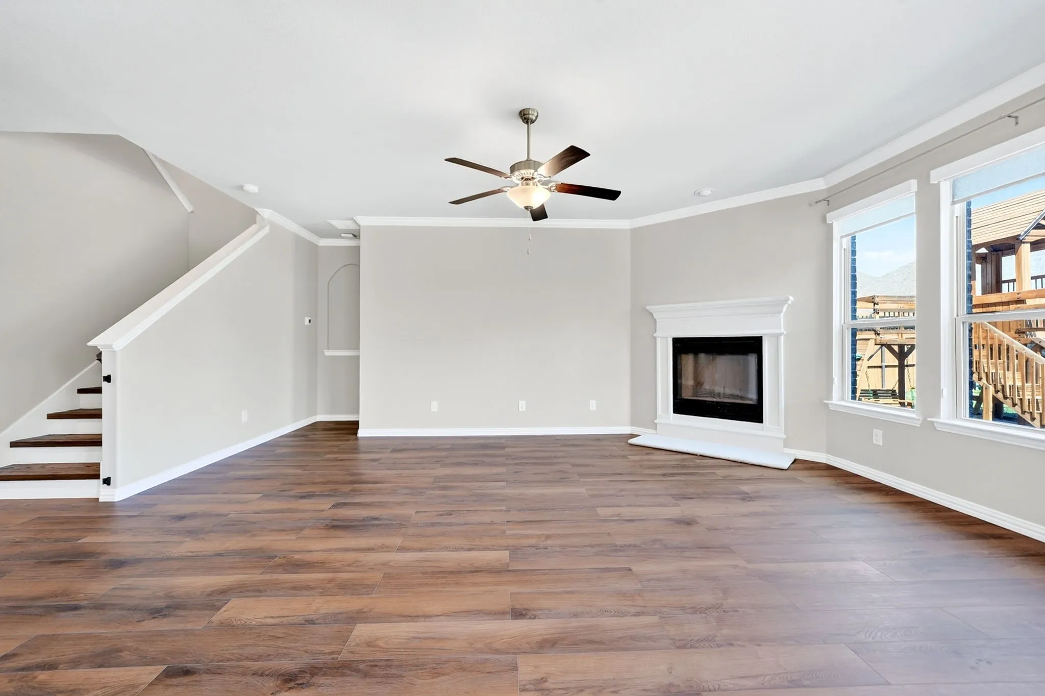 Spacious living area featuring wood-finish flooring, a white mantel fireplace, multiple windows, a ceiling fan, and staircase with dark treads and white risers