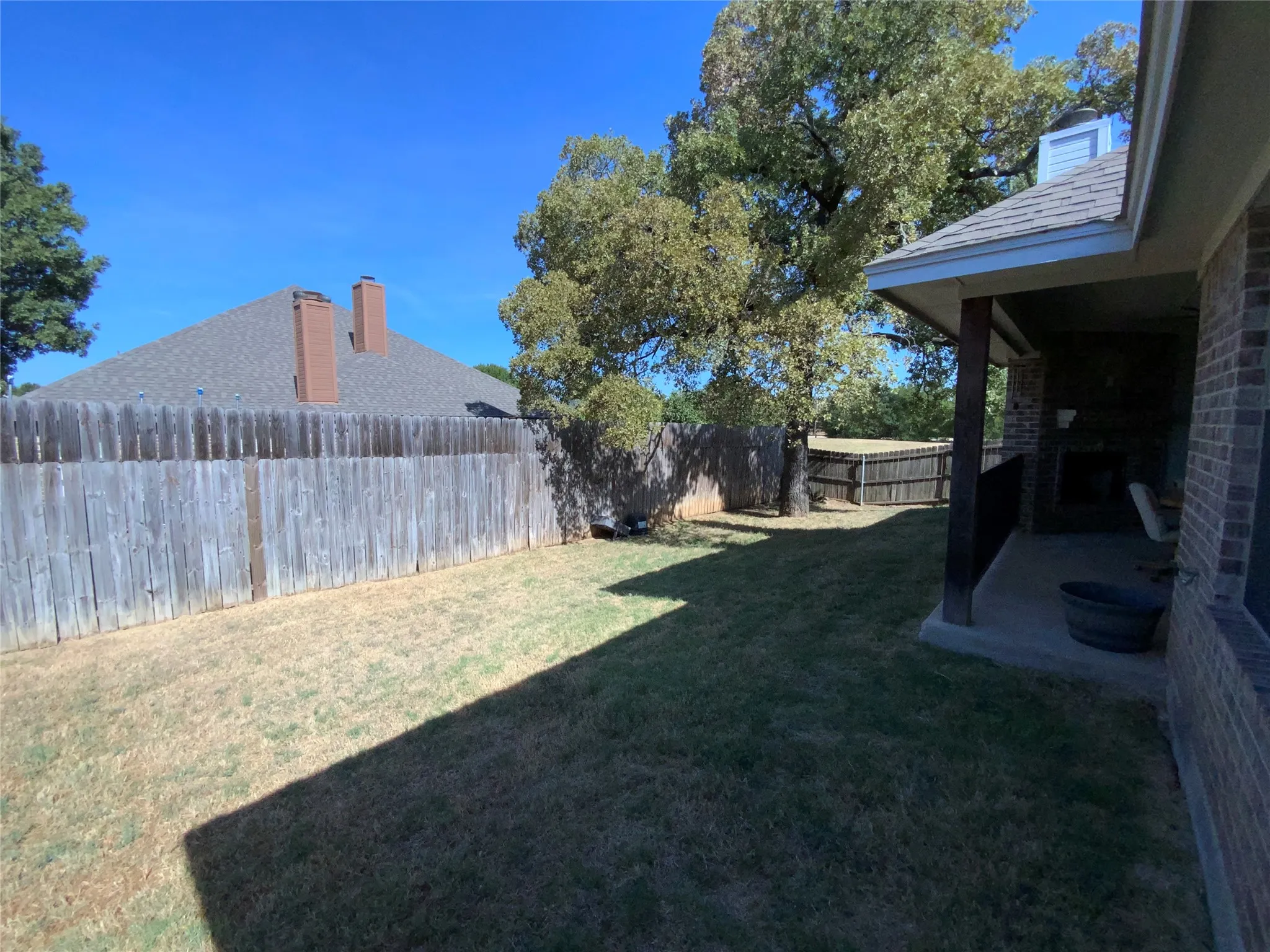 Expansive backyard featuring a wood fence, mature trees, and a covered patio with a brick exterior