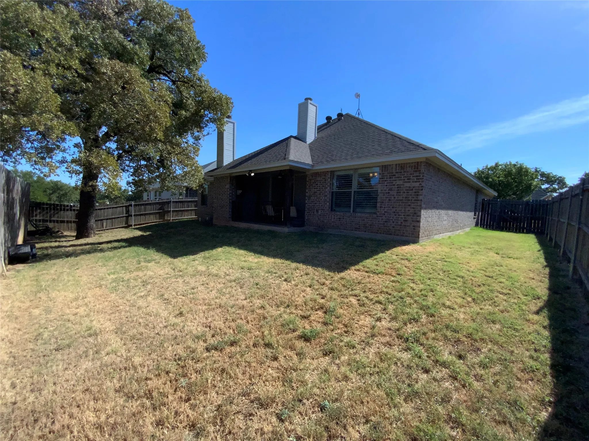 Enclosed backyard featuring a mature tree, brick exterior, covered patio, and privacy fencing