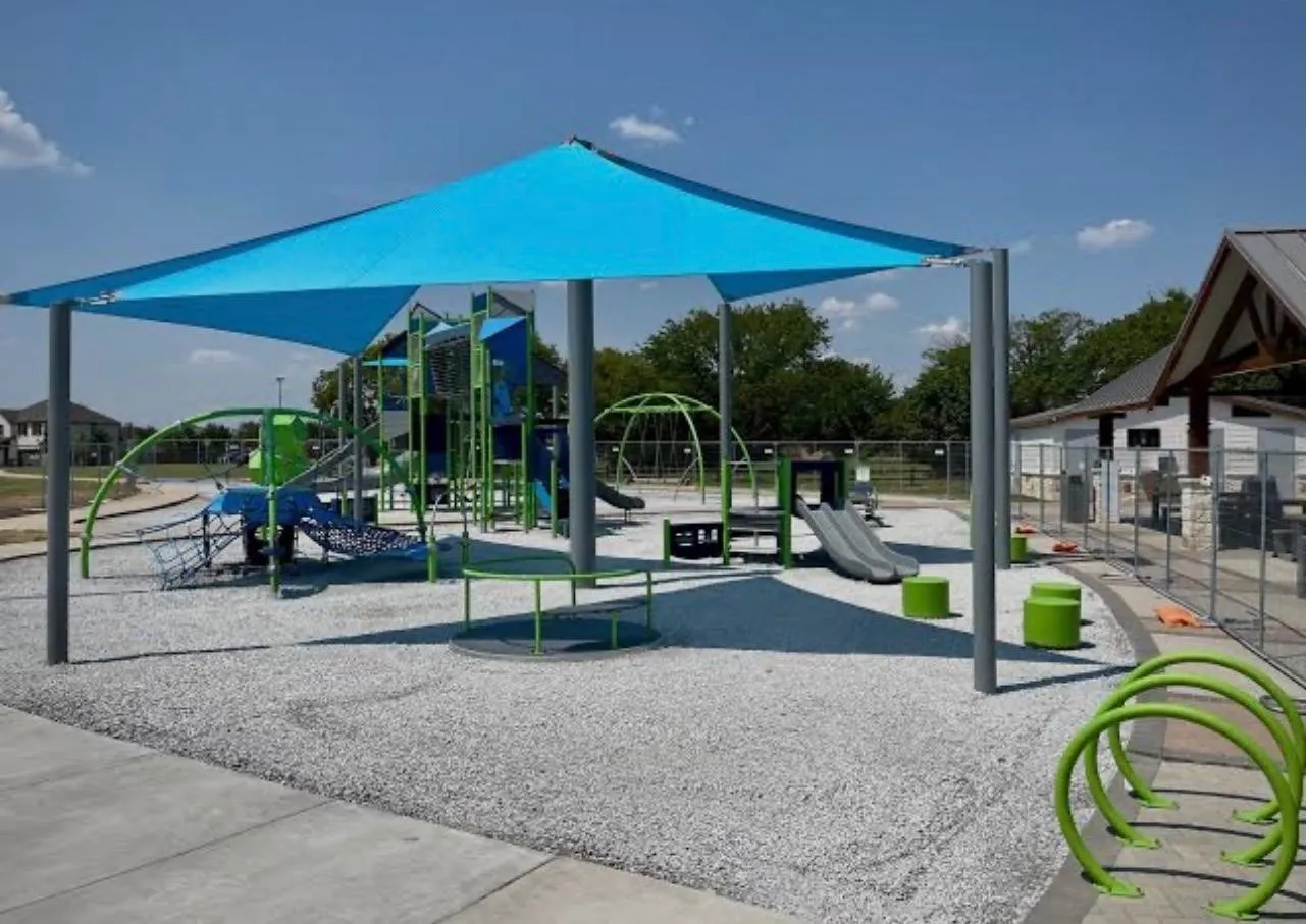 Playground featuring a blue shade structure, multiple play elements, a rubberized safety surface, and a nearby covered pavilion