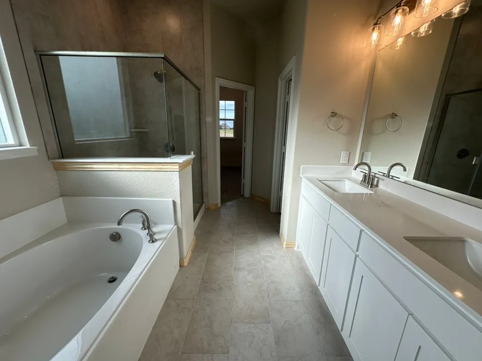 Dual sink vanity with white cabinetry and light-toned countertops, featuring a built-in tub with chrome fixtures, a glass-enclosed shower with tiled walls, and light-toned floor tiling