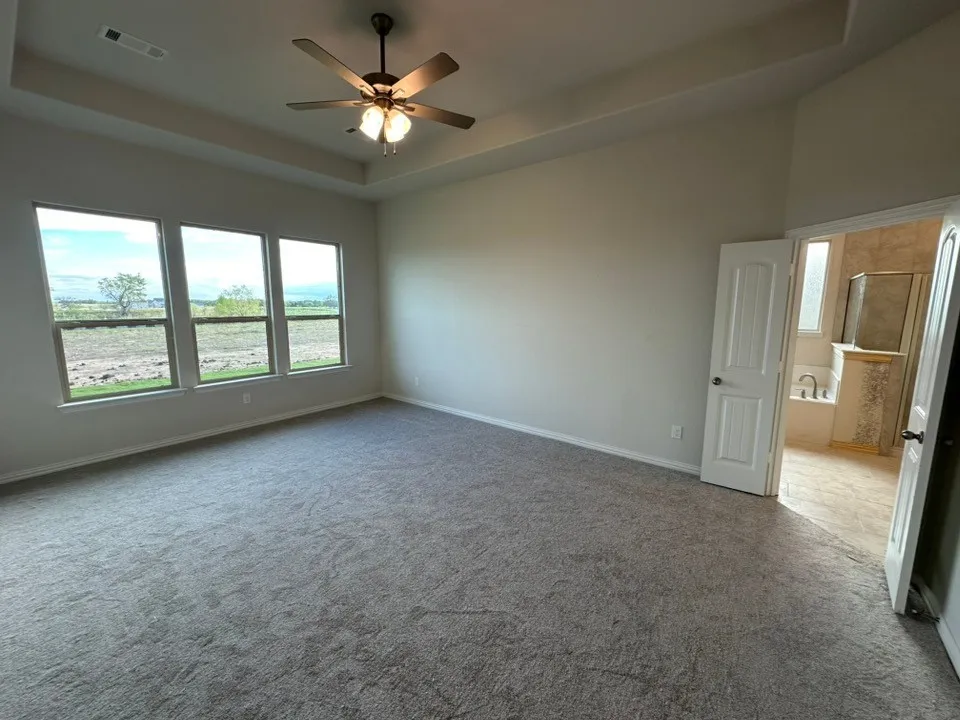 Tray ceiling with a ceiling fan, three pane windows, neutral carpet flooring, and an adjoining bathroom with a built-in tub