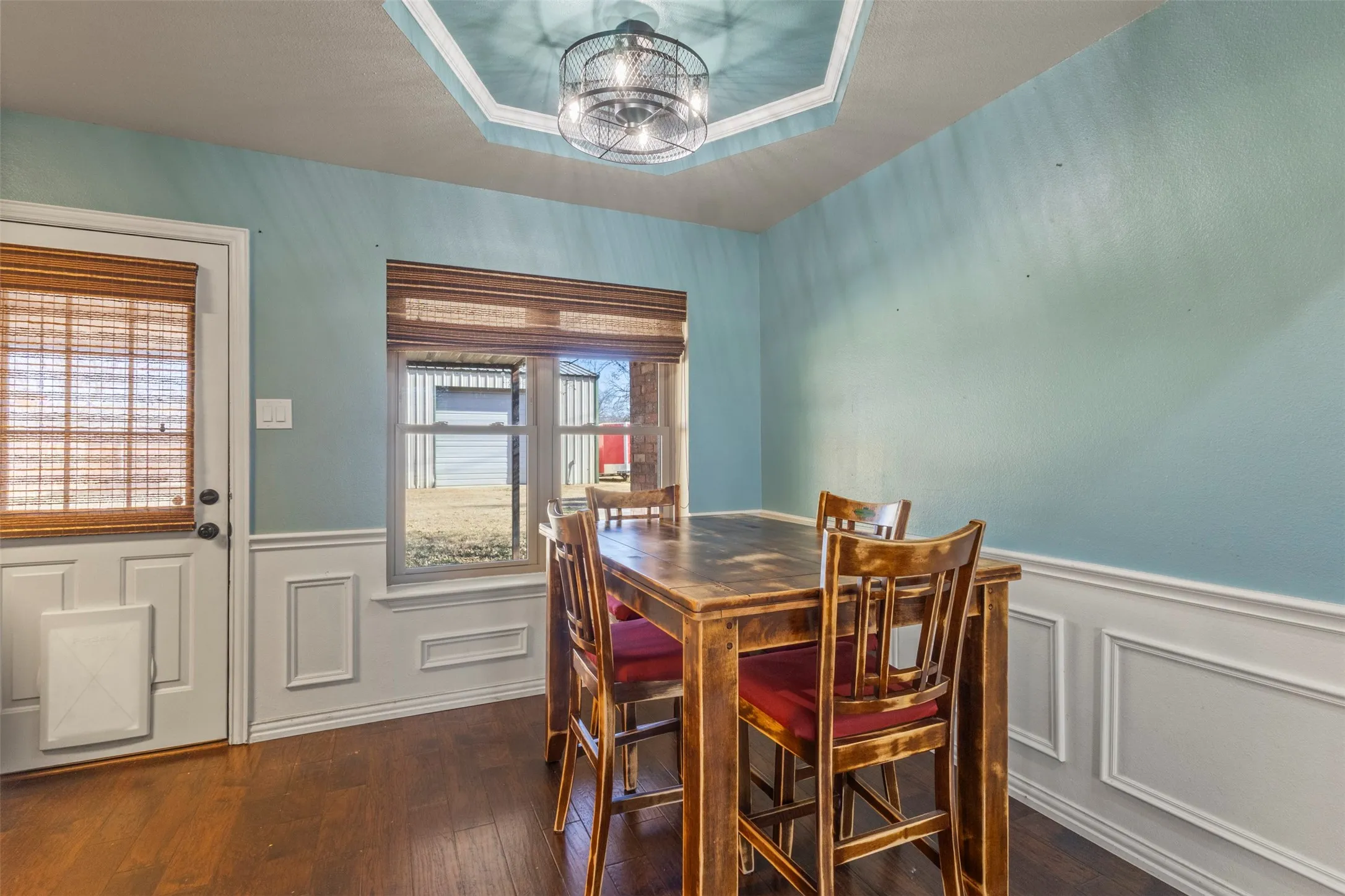 Tray ceiling with a decorative fan fixture, paneled wainscoting, wood-finish flooring, and a window with blinds