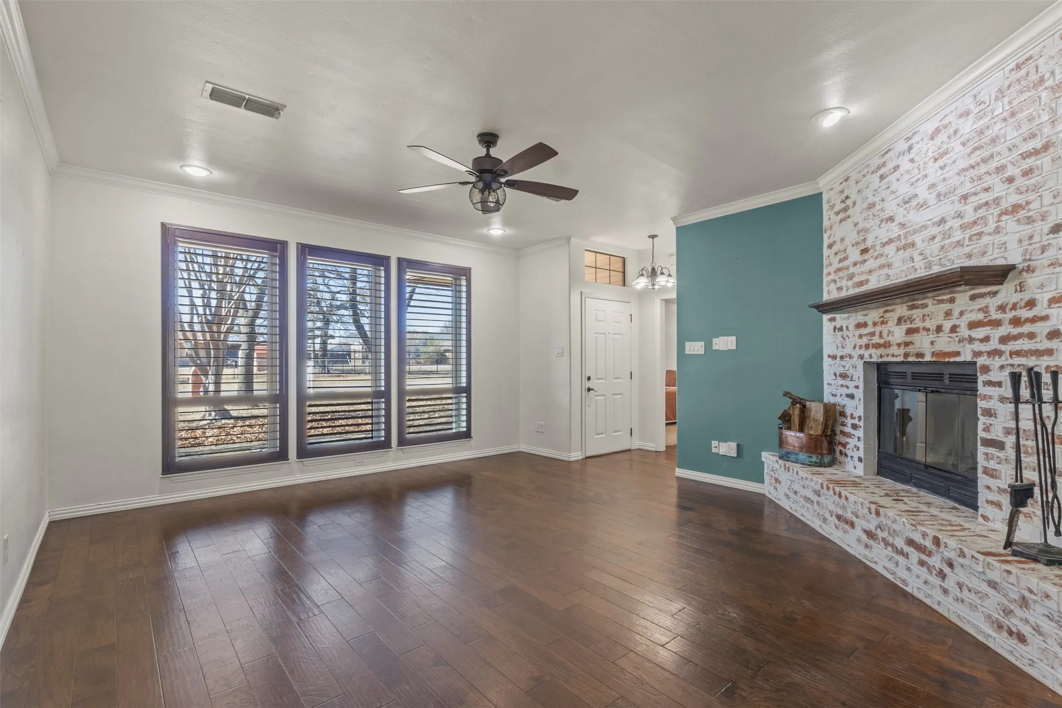 Wood-finish flooring, white brick fireplace with dark wood mantel, trio of windows with horizontal blinds, ceiling fan, and recessed lighting