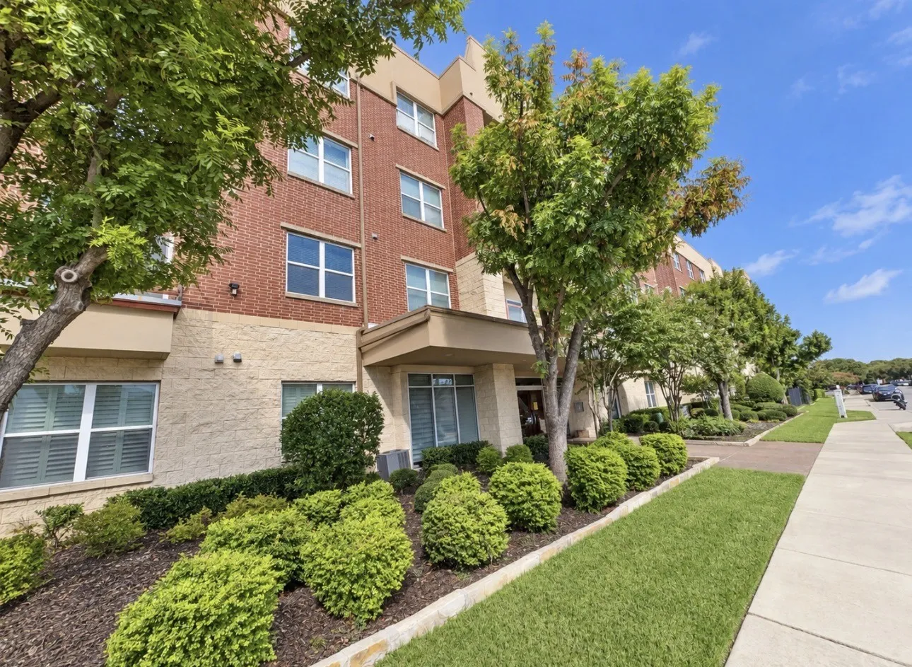 Multi-story building featuring brick and textured stucco siding, prominent entry portico, and mature landscaping with shrubs and trees