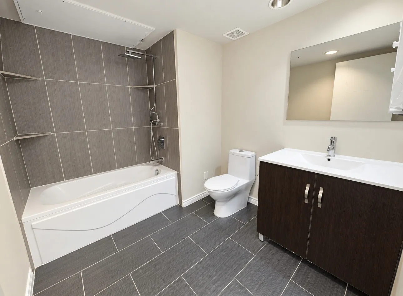 Bathroom featuring a white bathtub, gray tiled shower surround with built-in shelves, dark wood-finish vanity with an integrated sink, and large format gray tile flooring