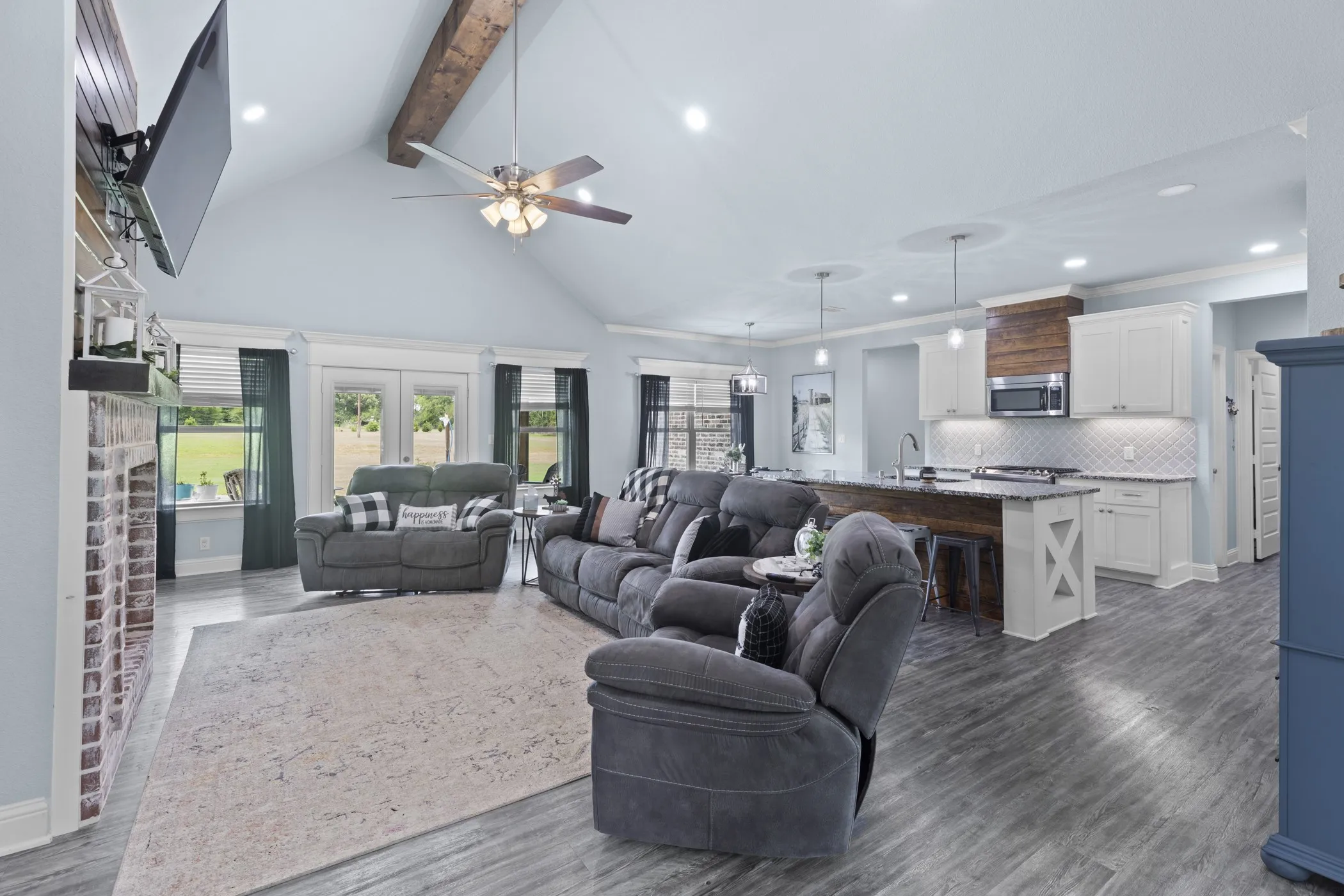Vaulted ceiling with exposed wood beam, wood-finish flooring, brick fireplace with mantel, white cabinetry, and a kitchen island with a wood-wrapped base