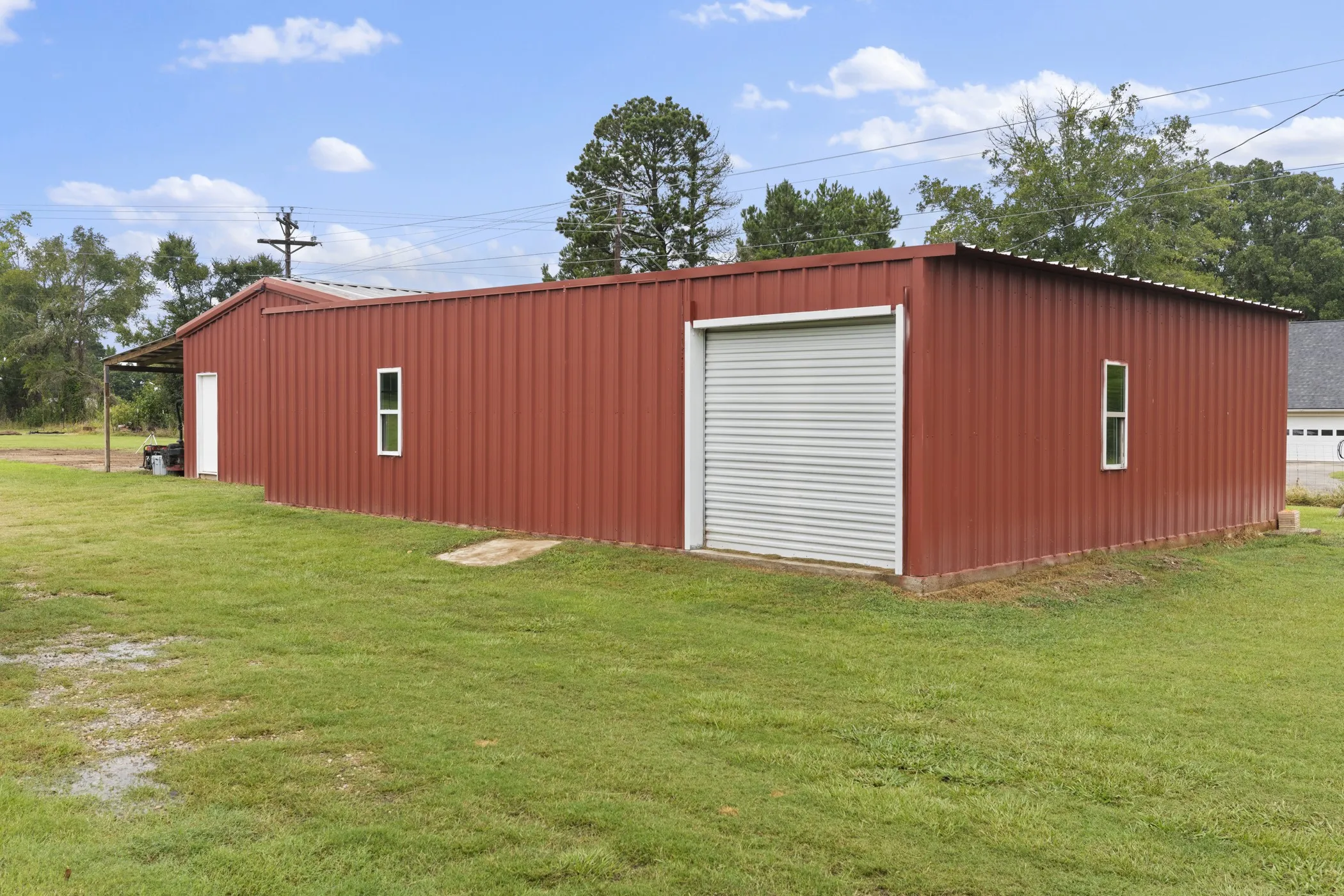 Red-colored metal building featuring a corrugated metal exterior, a roll-up garage door, white framed windows, a white entry door, and an attached covered extension
