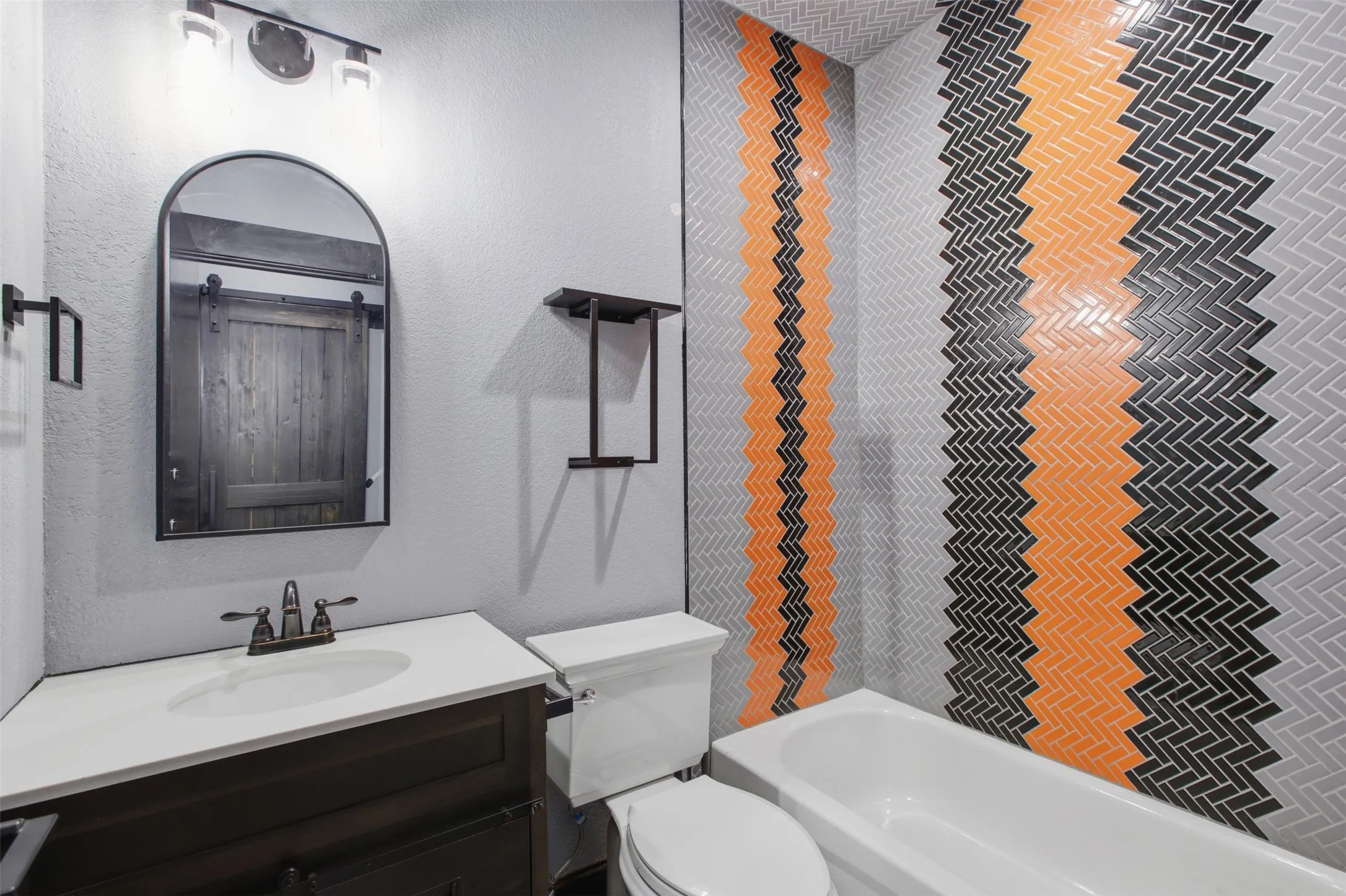 Herringbone tile wall with orange and black accents, white bathtub, dark wood-finish vanity with white countertop, arched mirror, and bronze-finish fixtures