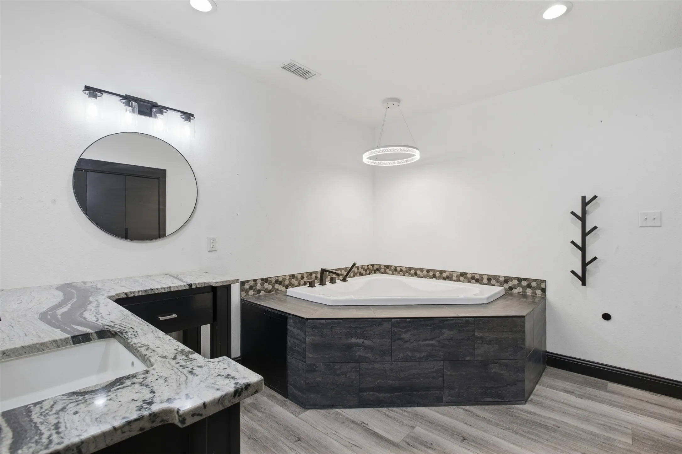 Spacious bathroom featuring a built-in hexagonal soaking tub with mosaic tile surround, dual vanity with undermount sinks and granite countertops, modern black vanity lighting, and wood-finish flooring