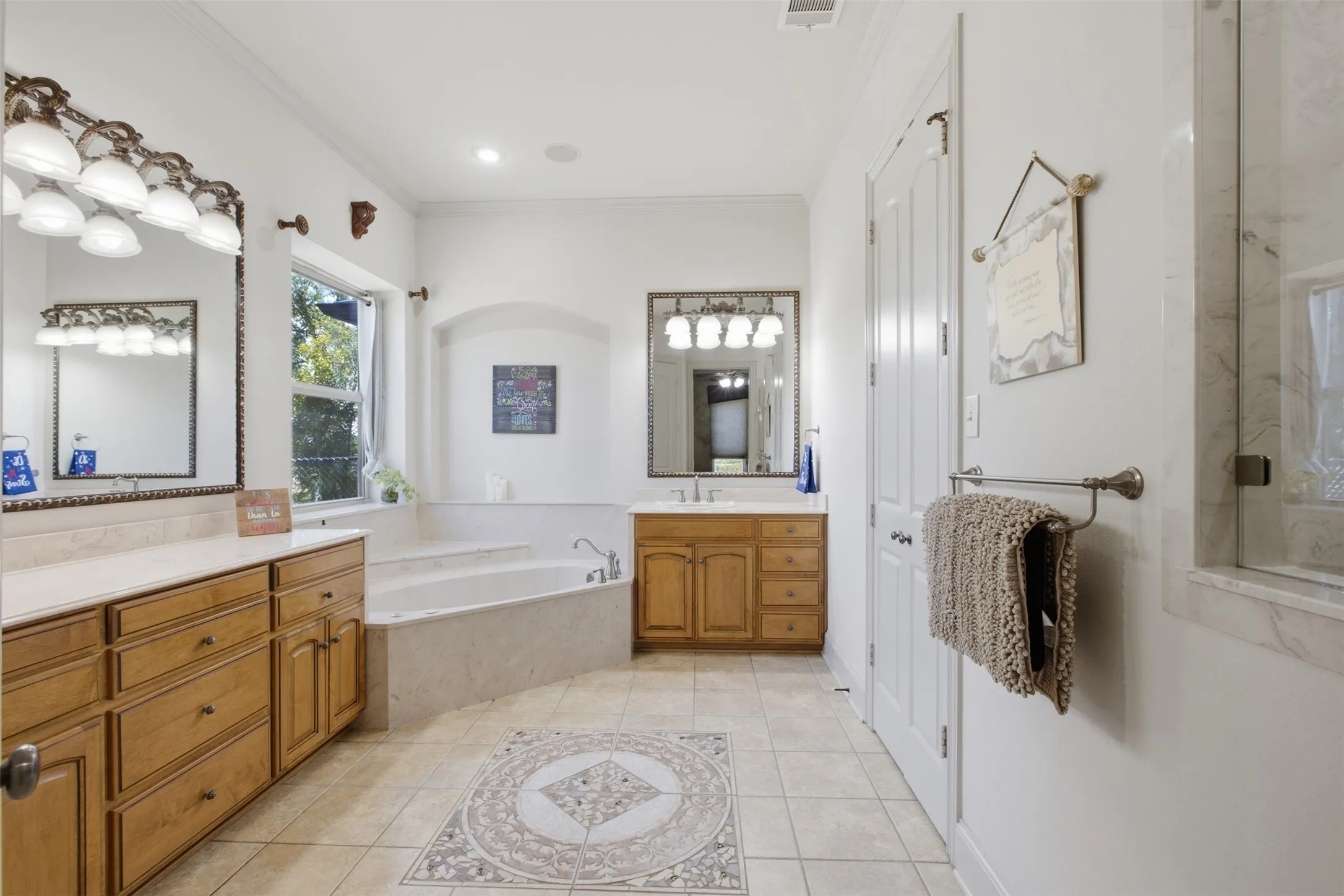 Expansive bathroom featuring dual wood-finish vanities with light countertops, a built-in soaking tub, tile flooring with an inset mosaic, and a glass-enclosed shower