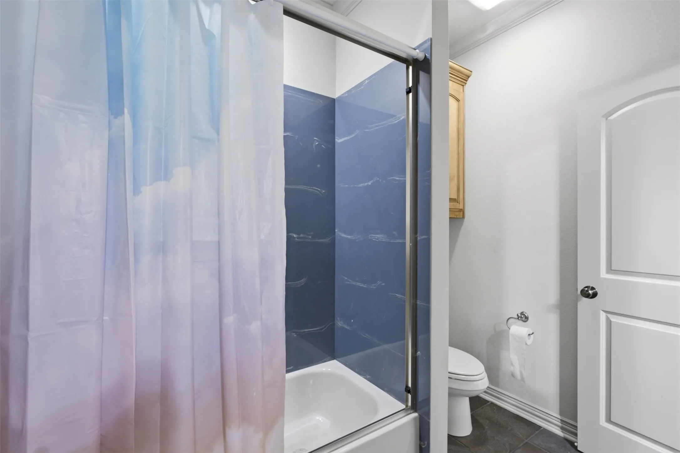 Bathtub with blue tiled surround, chrome shower enclosure, light wood-finish wall cabinet, white toilet, and gray floor tiles