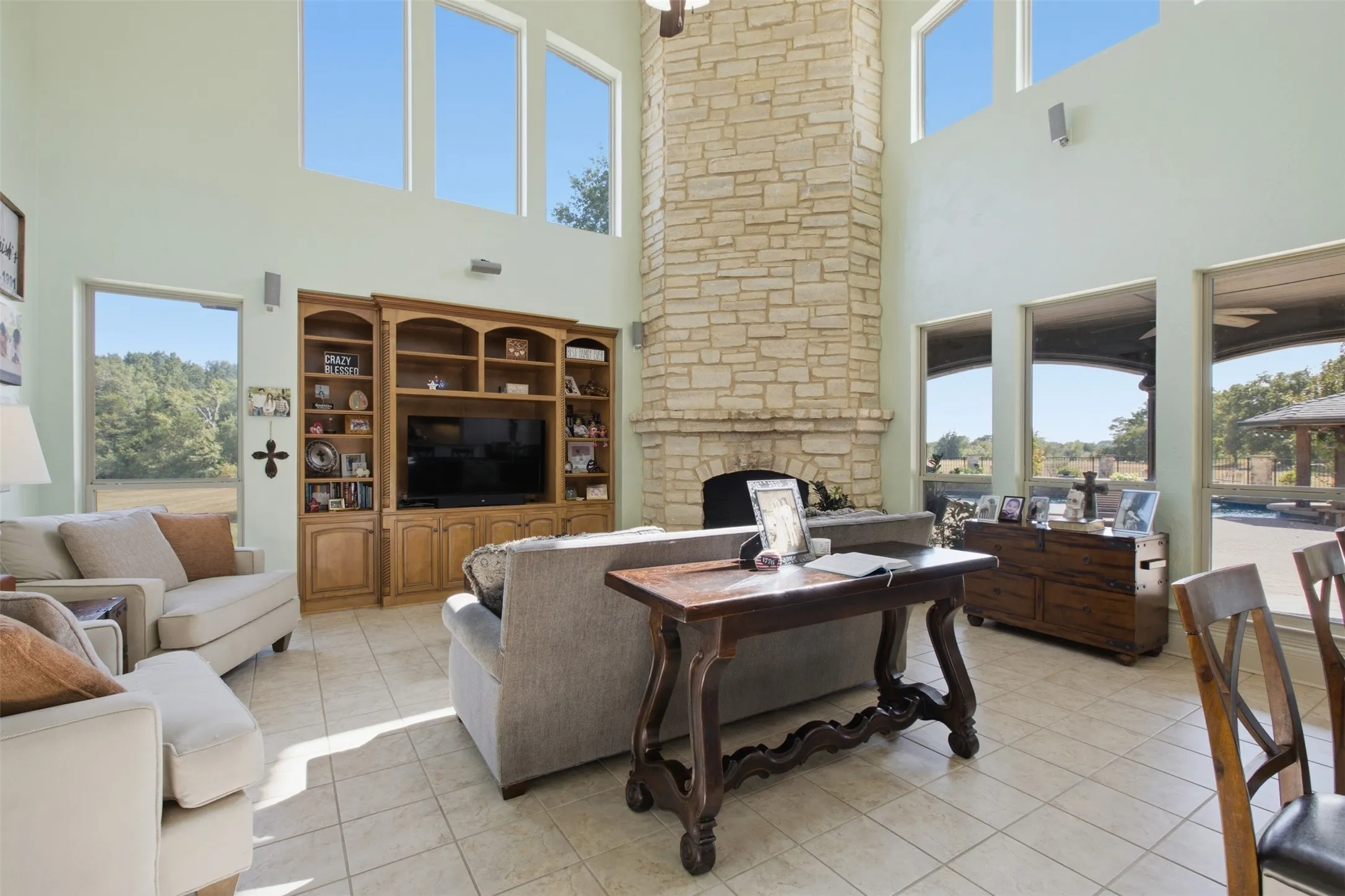 Two-story stone-clad fireplace with a raised hearth, built-in entertainment cabinetry, multiple large windows, and tile flooring