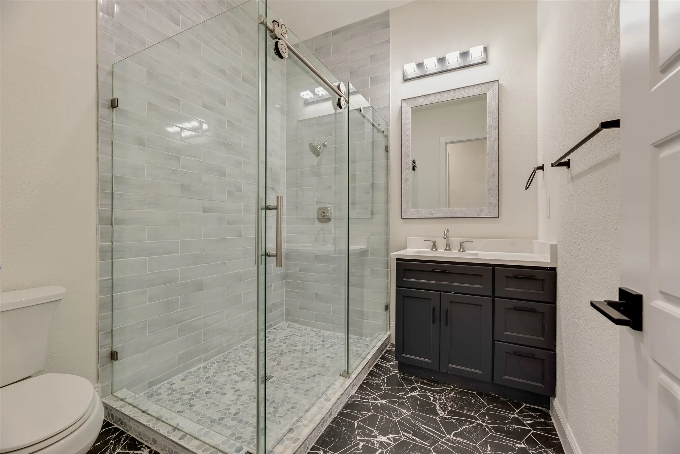Glass-enclosed shower with subway tile surround and pebble-style flooring, dark wood-finish vanity with countertop, and modern black geometric floor tiling