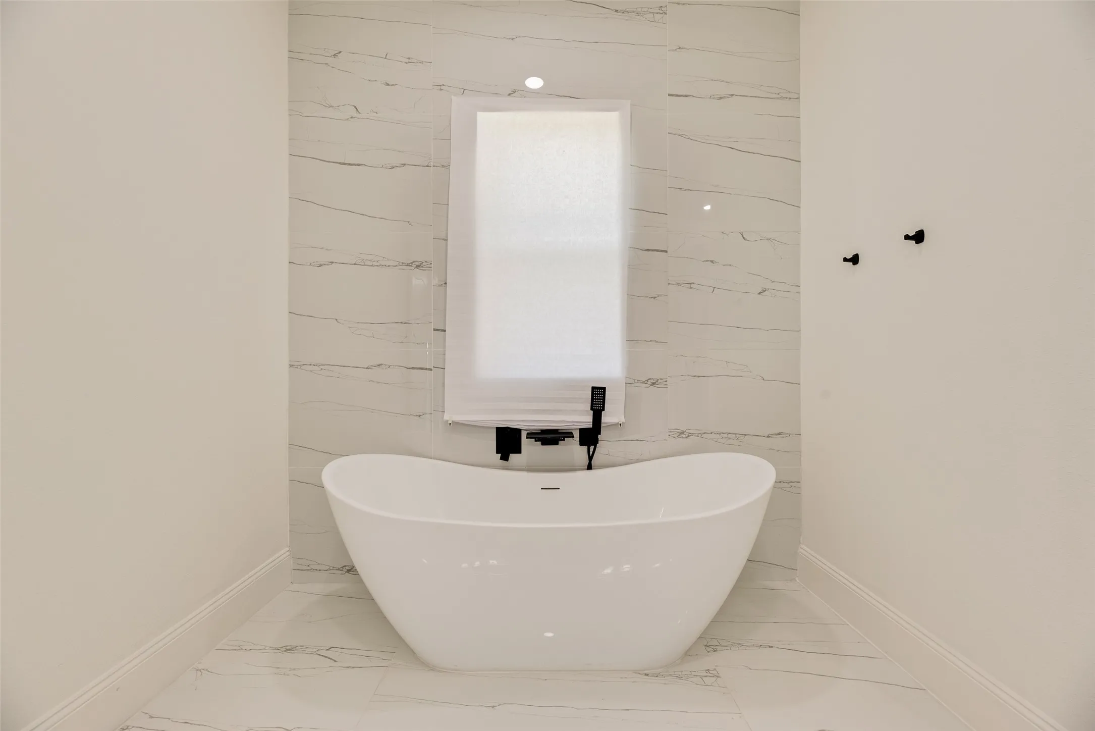 Freestanding soaking tub with a wall-mounted matte black faucet, set against a tiled accent wall featuring horizontal white tile with subtle veining, complemented by coordinated floor tile