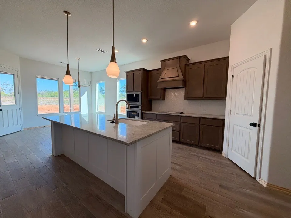 Spacious kitchen featuring a large island with a light-toned countertop, wood-finish flooring, dark wood cabinetry with a custom vent hood, and a geometric tiled backsplash